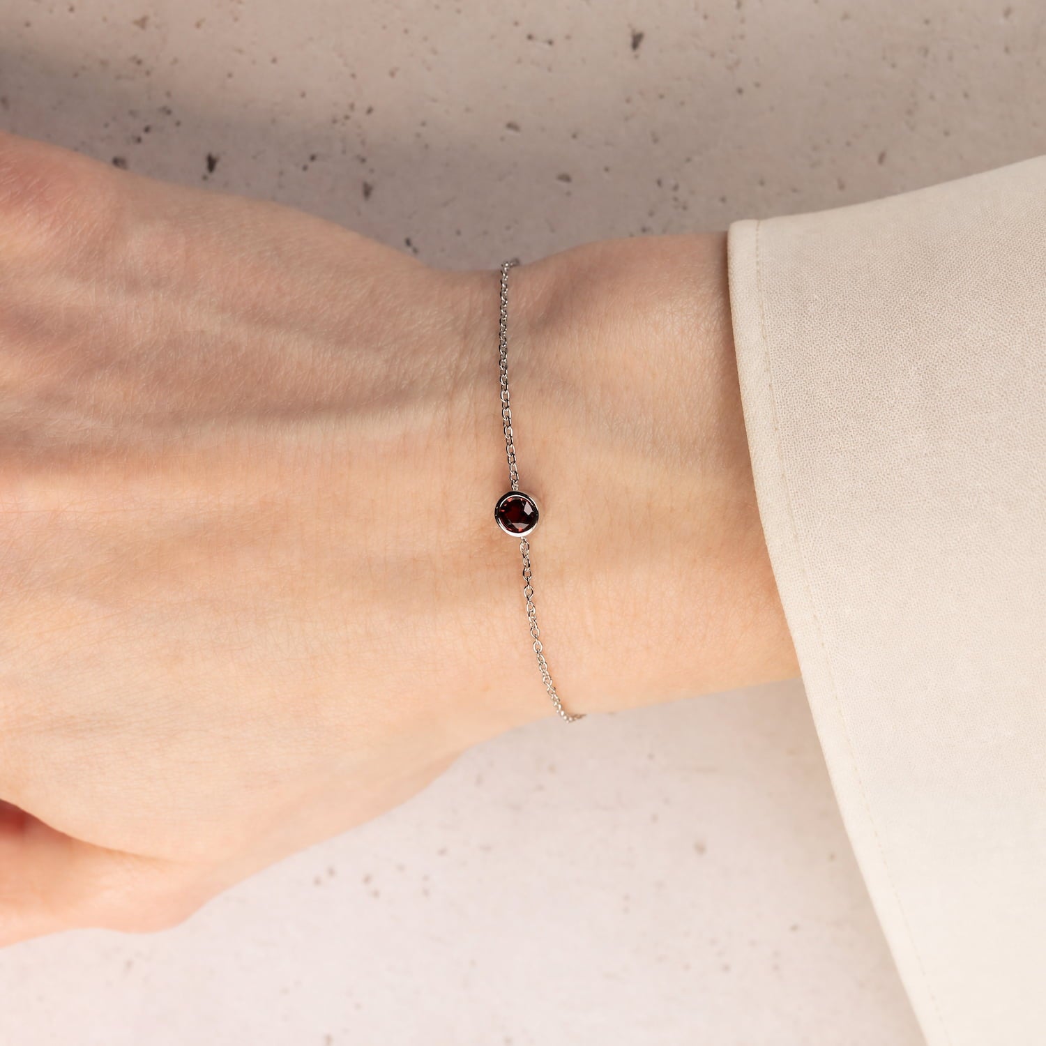 Delicate sterling silver bracelet featuring a deep red garnet birthstone set in a minimalist bezel, resting on a light neutral surface for a contemporary and elegant look.