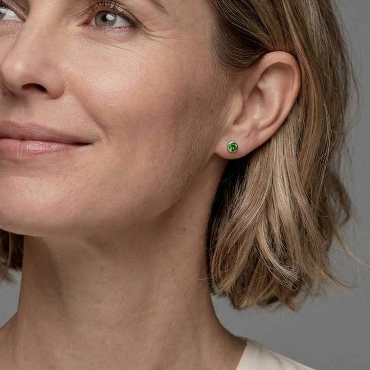 Elegant sterling silver stud earrings featuring vibrant Siberian emerald gemstones, worn by a woman with short blonde hair against a neutral grey background.