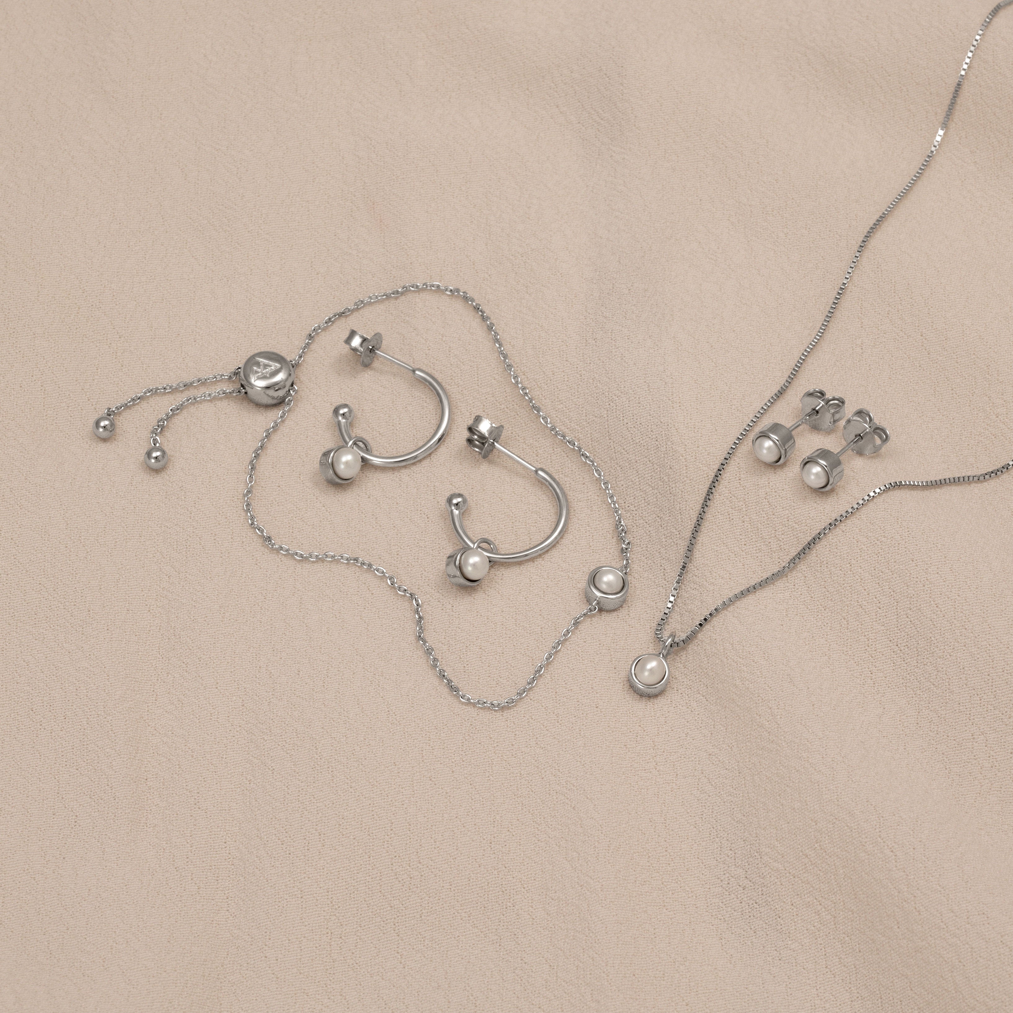 Collection of sterling silver jewellery featuring freshwater pearl accents including hoop earrings, stud earrings, a delicate pendant on a fine chain, and an adjustable bracelet displayed on a soft taupe fabric.