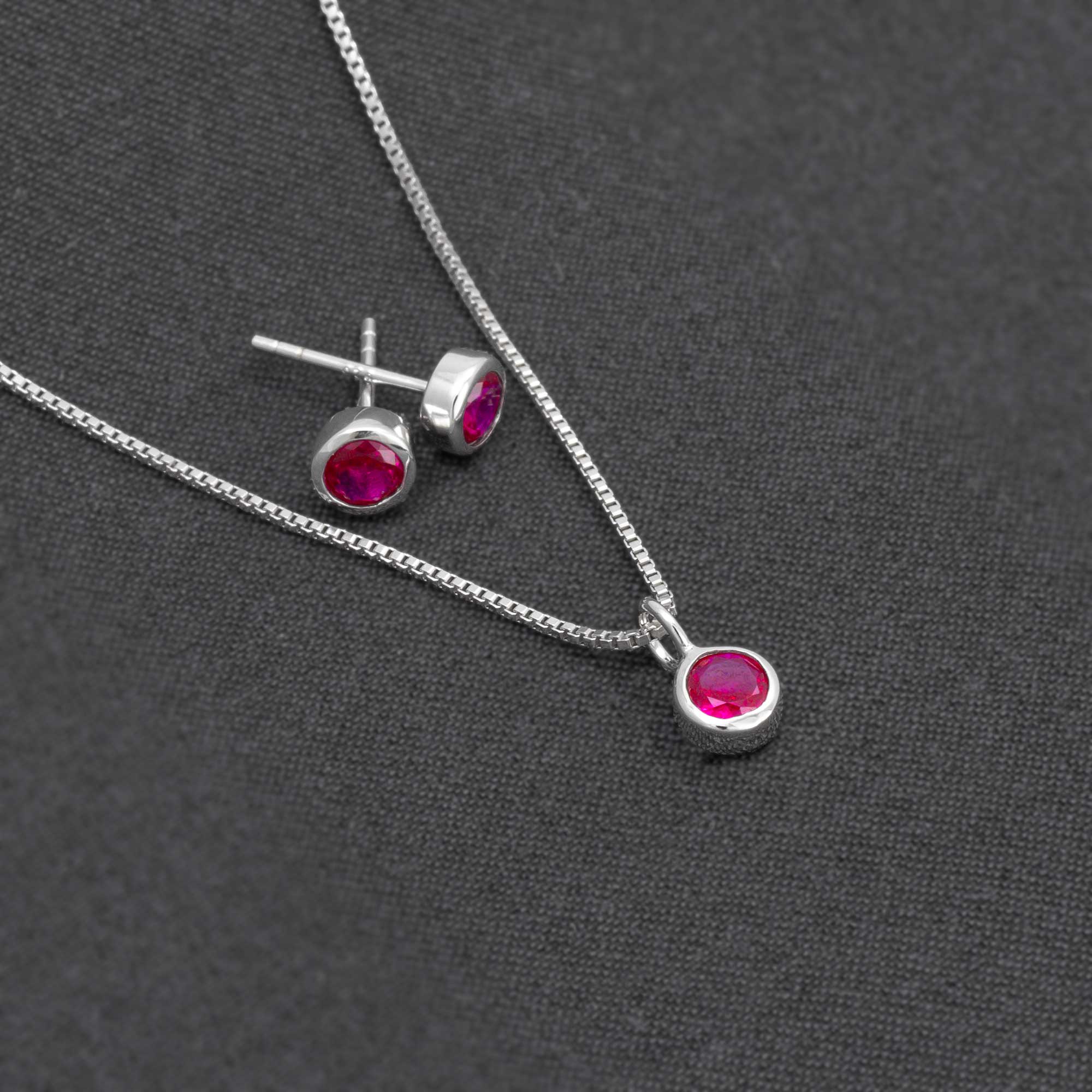 Pair of ruby gemstone stud earrings in sterling silver, displayed on a dark fabric background alongside a matching pendant with a fine silver chain, highlighting expert British craftsmanship.