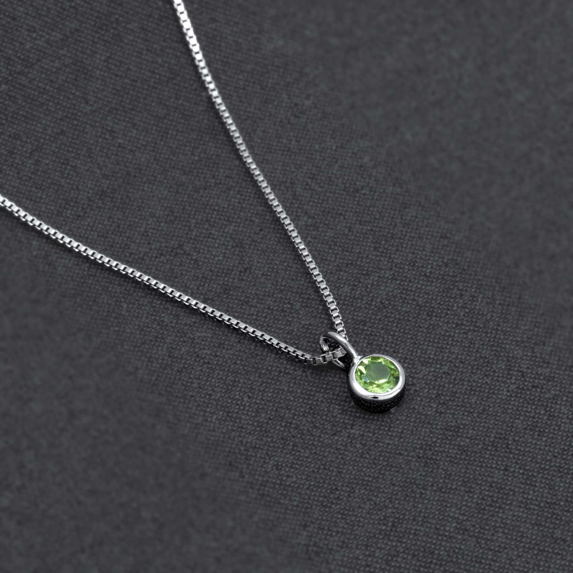 Delicate sterling silver pendant featuring a bezel-set peridot gemstone, suspended on a fine chain against a dark textured background, showcasing refined craftsmanship.