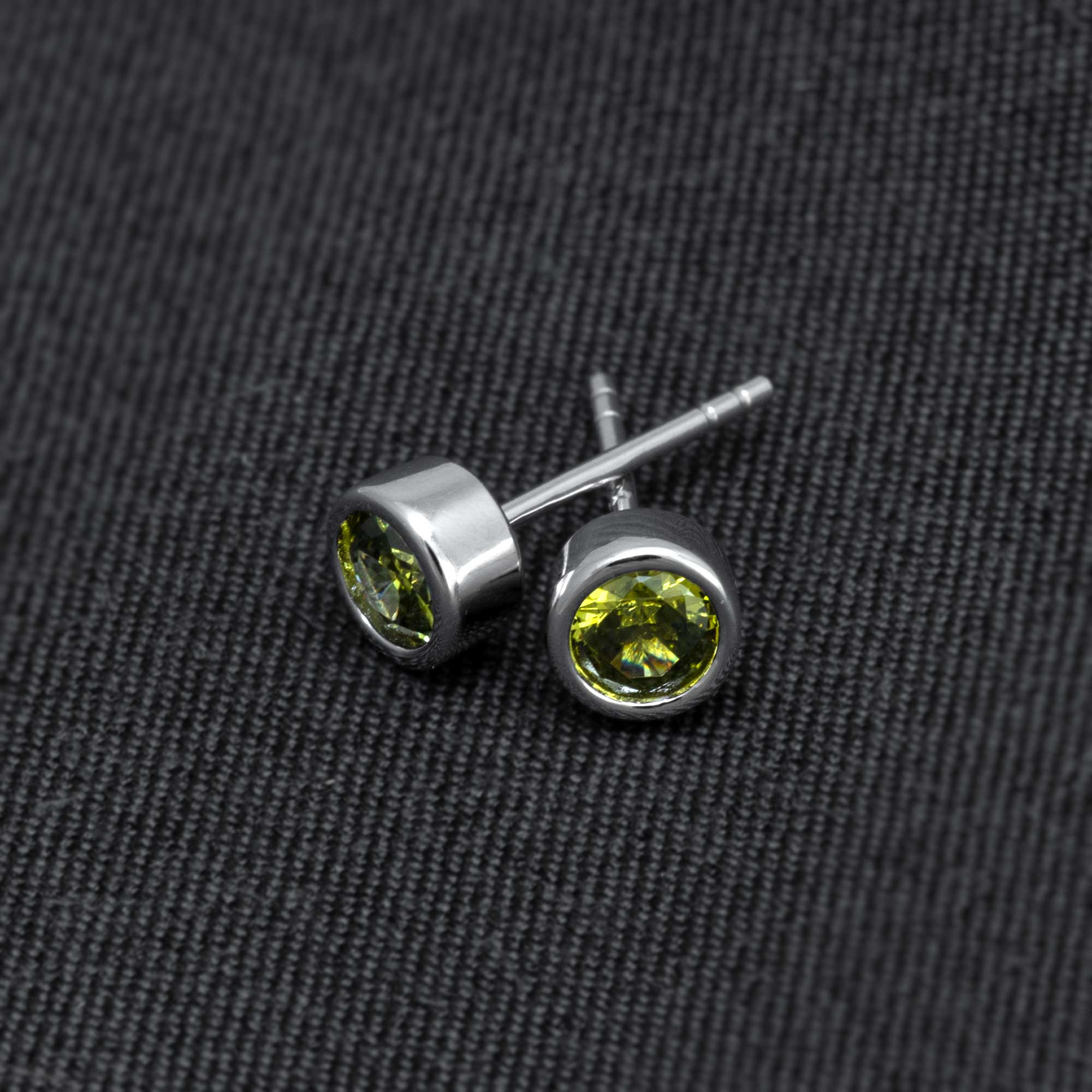Pair of sterling silver stud earrings featuring vibrant green peridot gemstones set in a sleek, contemporary bezel design on a textured dark fabric background.