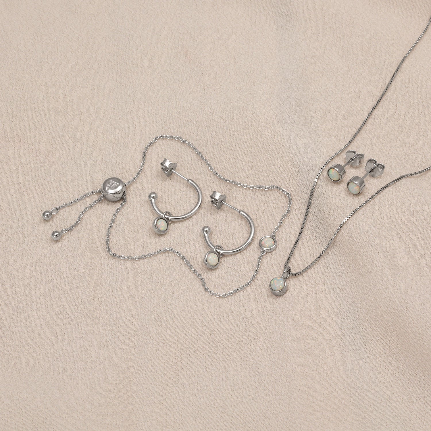 Delicate sterling silver jewellery set featuring opal stones, including hoop earrings, studs, a pendant necklace, and an adjustable bracelet, styled on a neutral fabric.