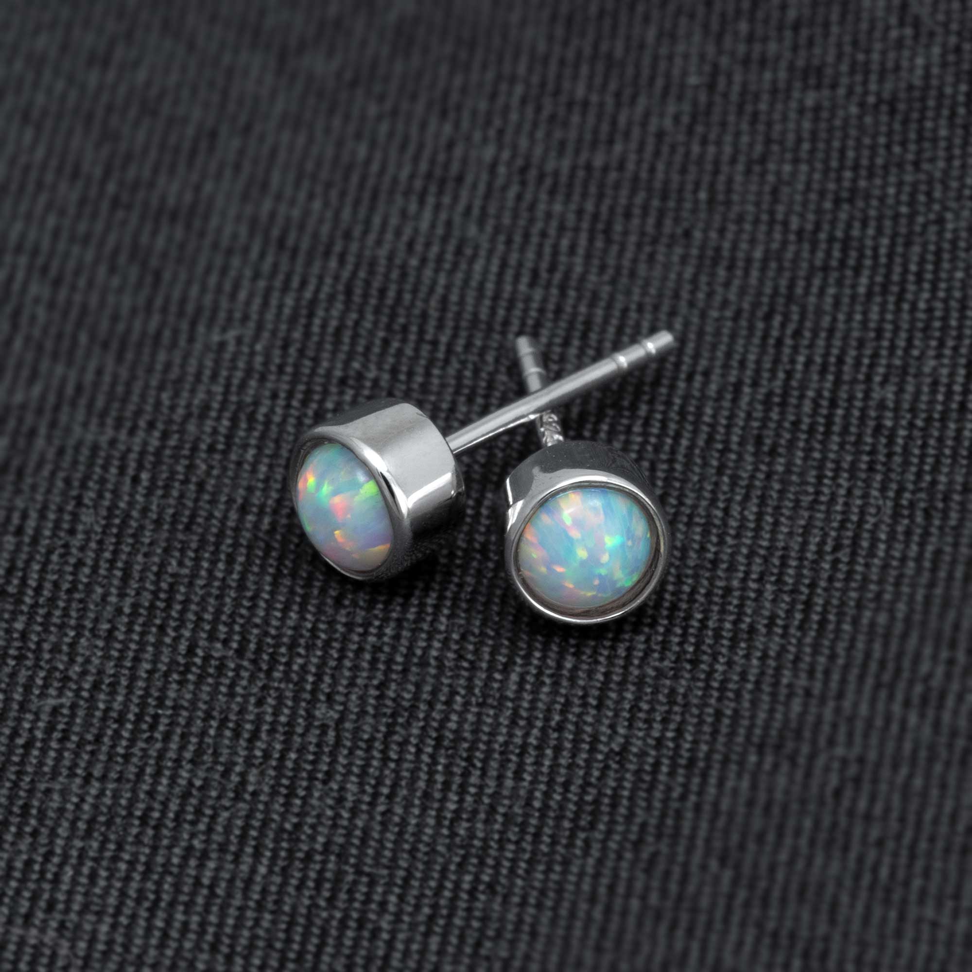 Pair of sterling silver stud earrings featuring round opal gemstones with a polished bezel setting, highlighting the iridescent play of colors against a dark fabric background.