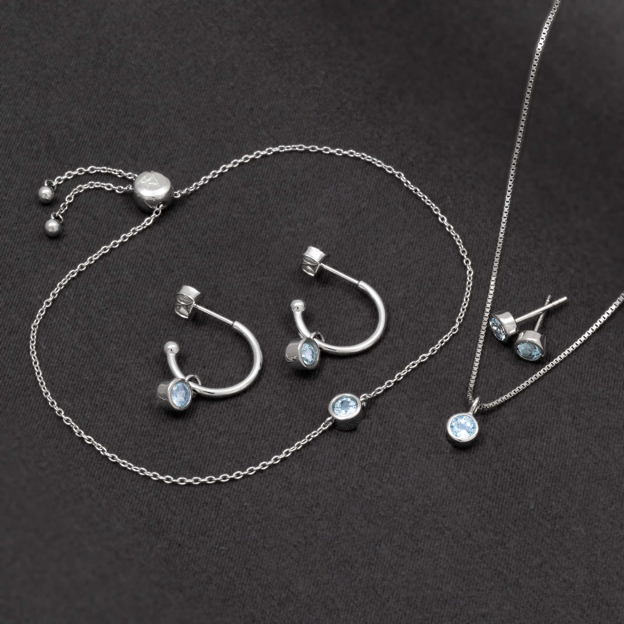 Sterling silver hoop earrings and stud earrings featuring round blue topaz gemstones, accompanied by a delicate matching bracelet and pendant on a fine chain.