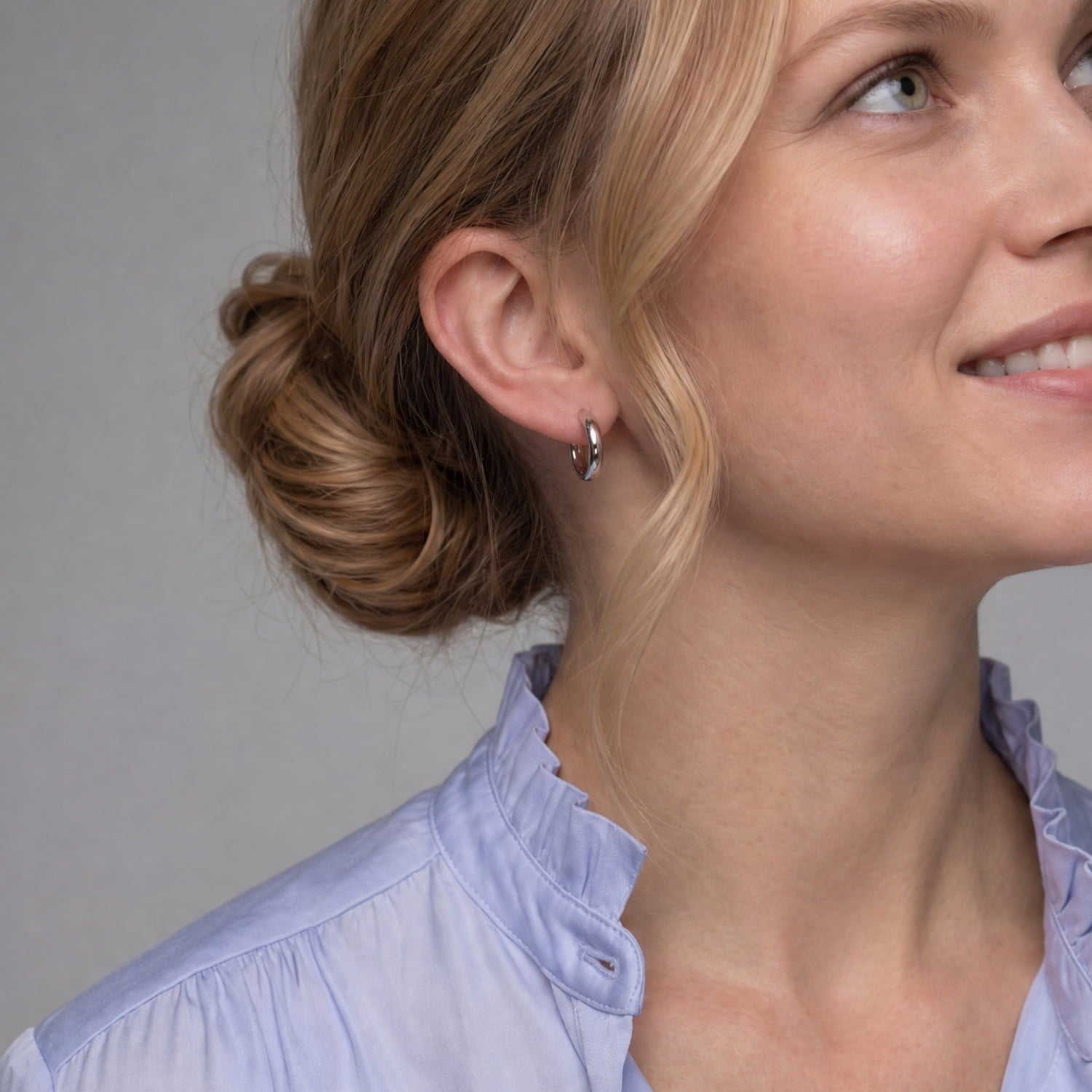 A close-up of a woman wearing a small silver hoop earring, with her hair in a bun and a light blue blouse against a plain gray background.