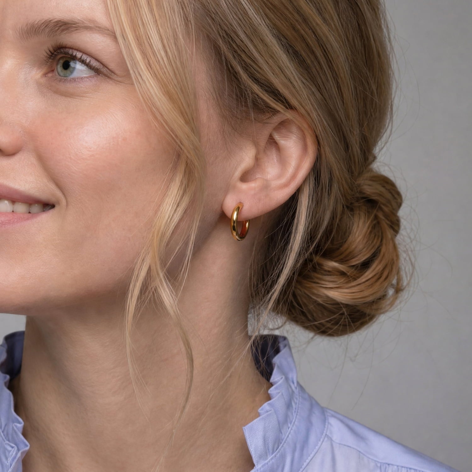 A close-up of a woman wearing a small gold vermeil hoop earring, with her hair in a low bun and a light blue ruffled shirt visible against a plain gray background.