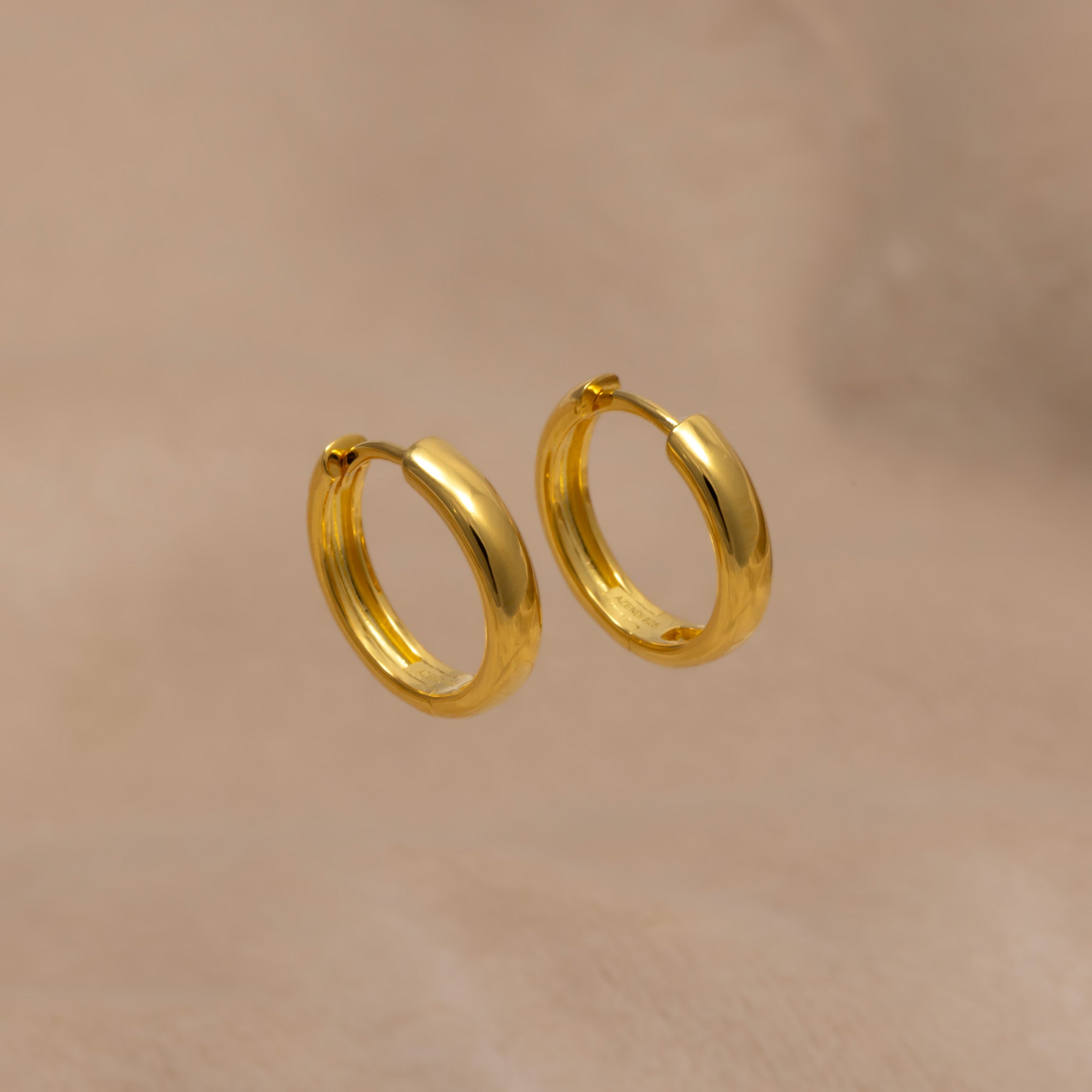 Pair of medium rounded hinge hoop earrings in polished gold vermeil, showcasing a sleek, classic design perfect for everyday elegance in fine jewellery. Placed on a cream velvet cloth.