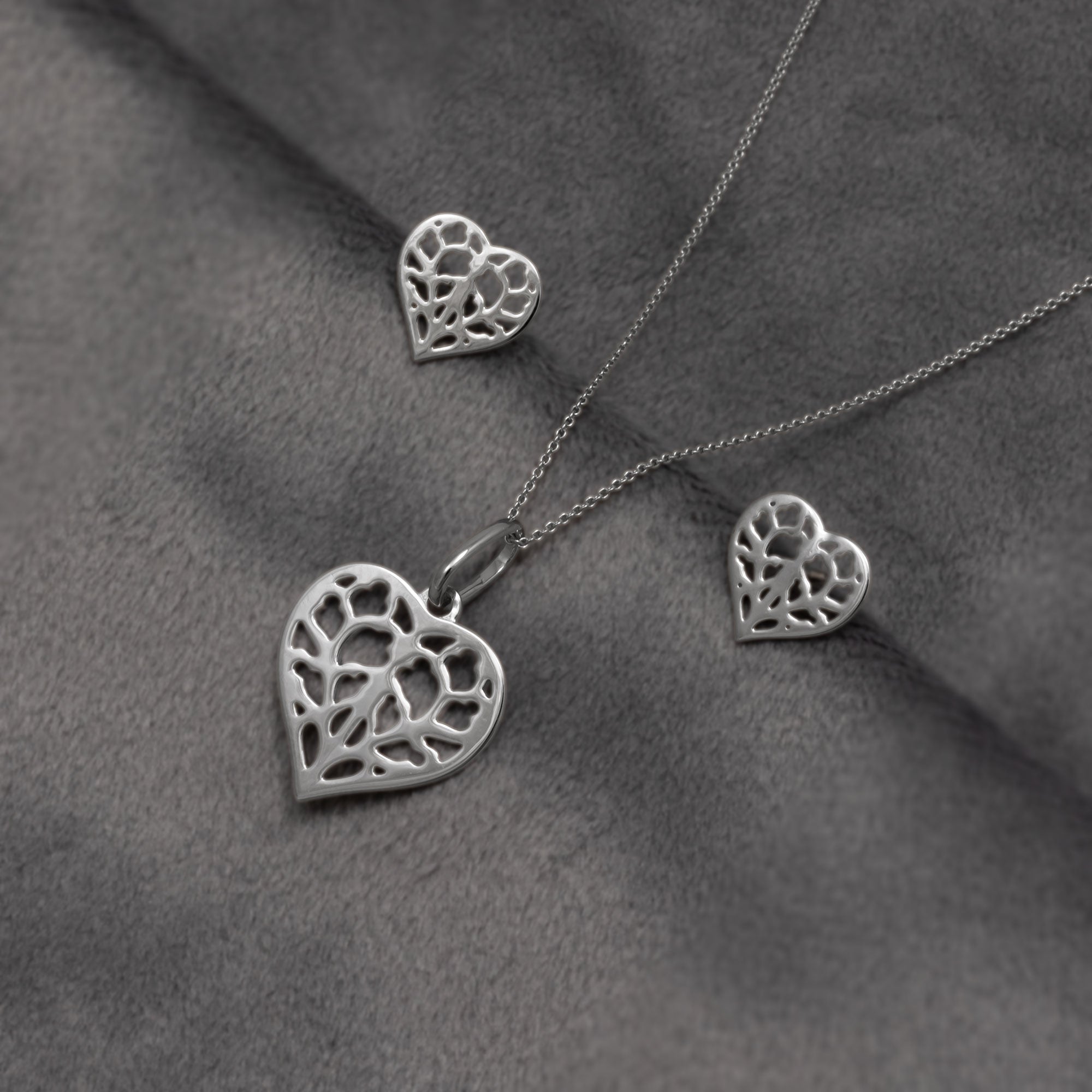 Pair of 9 carat white gold stud earrings and matching pendant featuring delicate openwork heart shapes inspired by York Minster, combining polished surfaces with a refined sculptural design. Placed on a grey velvet cloth.