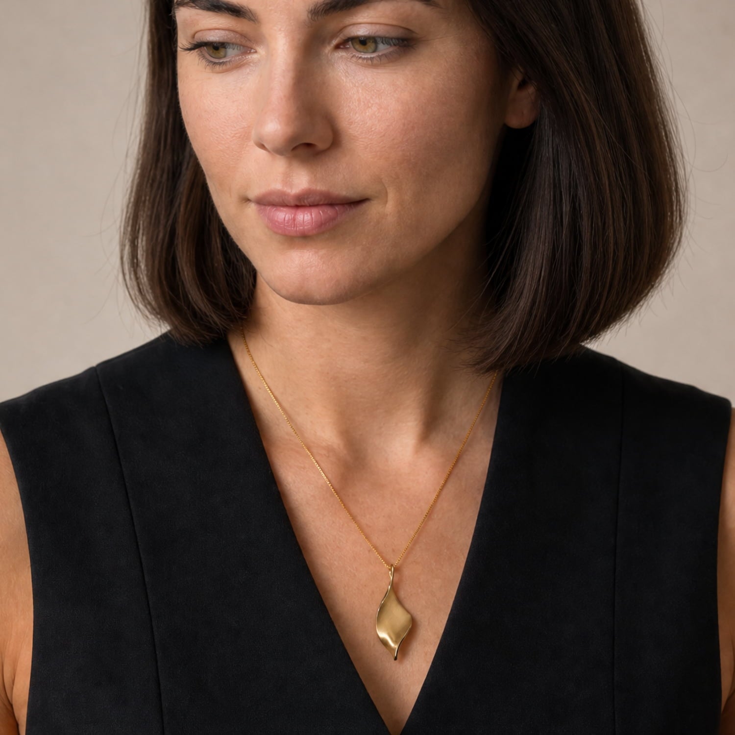 A close-up of a woman with dark brown shoulder-length hair wearing a gold vermeil twisting pendant necklace and a black sleeveless top, set against a neutral beige background.
