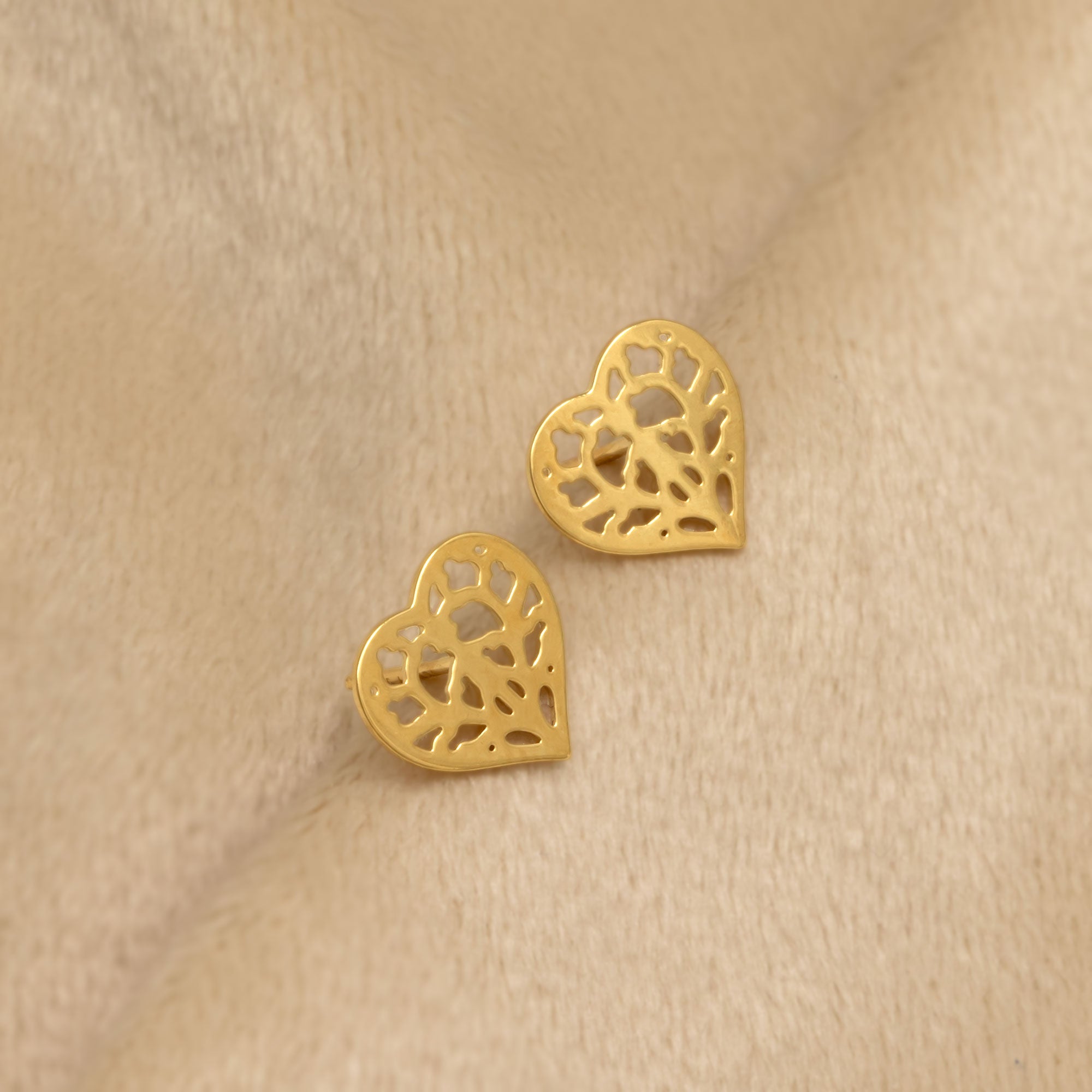 Pair of 9 carat yellow gold stud earrings featuring an intricate heart-shaped openwork design, combining delicate artistry with a refined contemporary finish. Placed on a cream velvet cloth.