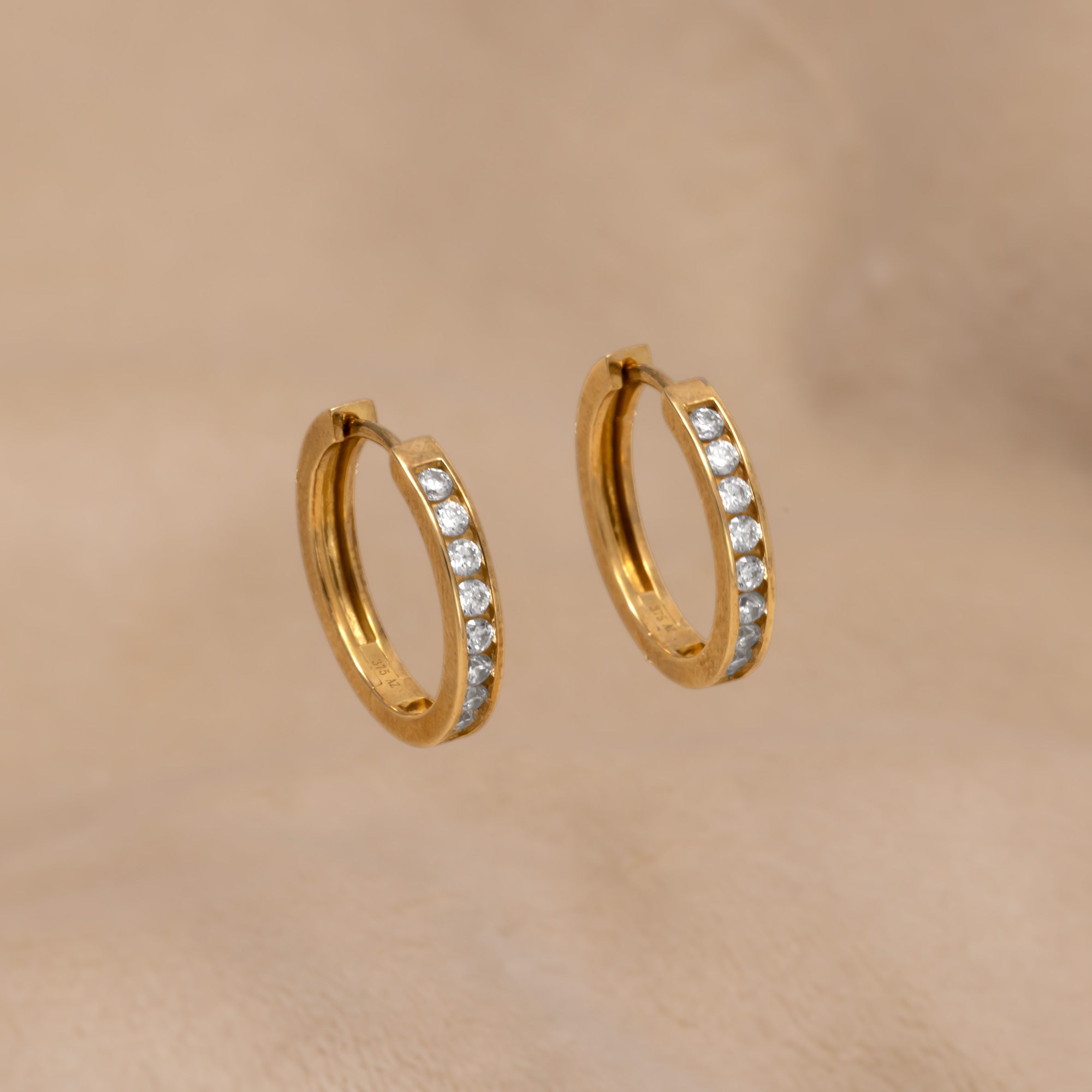 Pair of 9 carat gold hinge hoop earrings featuring a row of medium, clear stones for a refined and sculptural accessory with a modern elegance. Placed on a cream velvet cloth.