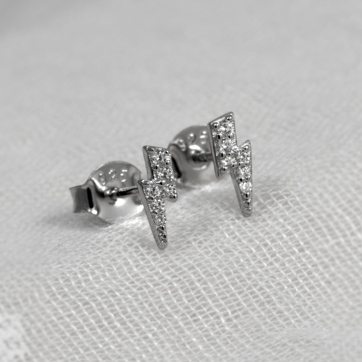 A pair of silver lightning bolt stud earrings set with small clear stones, displayed on a textured white fabric background.