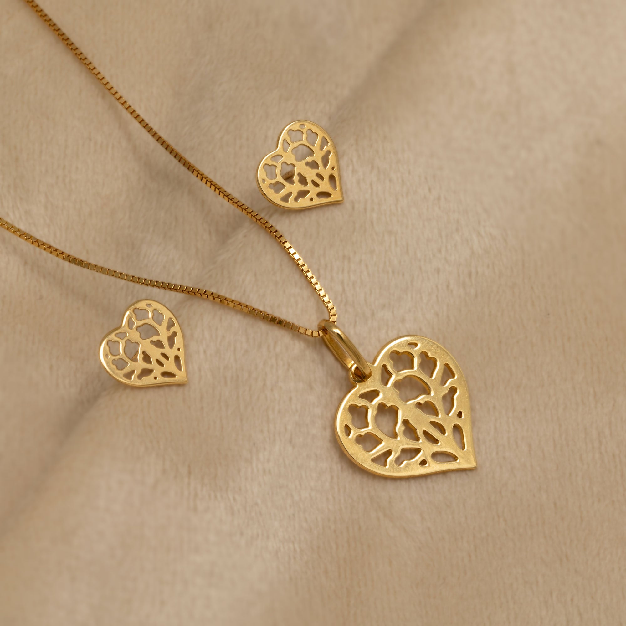 An 18ct yellow gold heart-shaped pendant and matching stud earriings featuring an intricate openwork design inspired by York Minster. It is suspended from a delicate chain for an elegant and sculptural appearance. Placed on a cream velvet cloth.