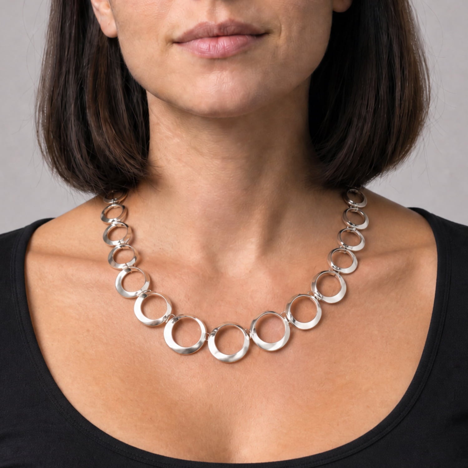 A close-up of a woman wearing a chunky silver necklace made of linked open rings, paired with a black scoop-neck top against a neutral gray background.