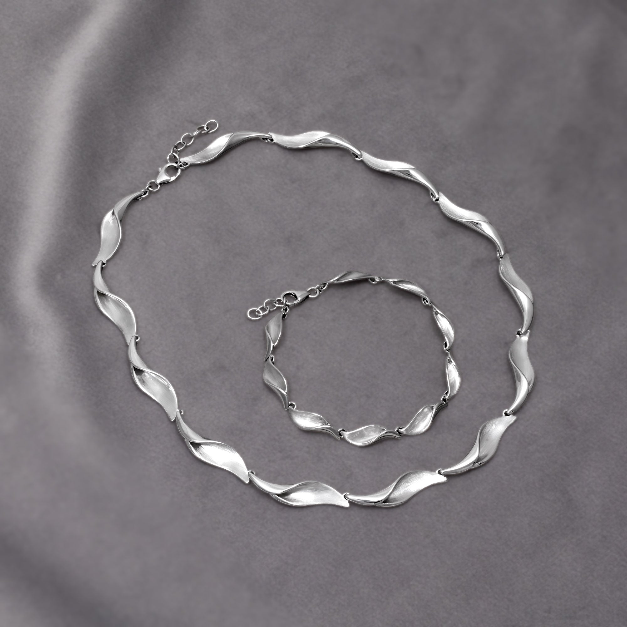 Sterling silver necklace and matching bracelet featuring alternating satin and polished links shaped as folded waves, creating a fluid sculptural effect for contemporary silver jewellery. Placed on a grey velvet cloth.