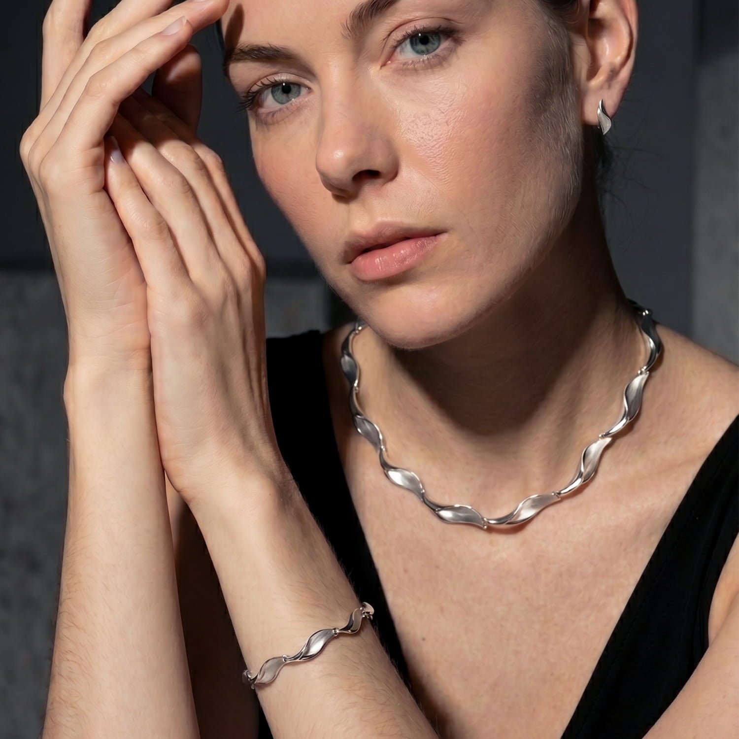 A close-up lifestyle shot of a woman wearing a matching silver twisted-link jewellery set, including a wavy chain necklace and a matching bracelet, against a dark gray background.