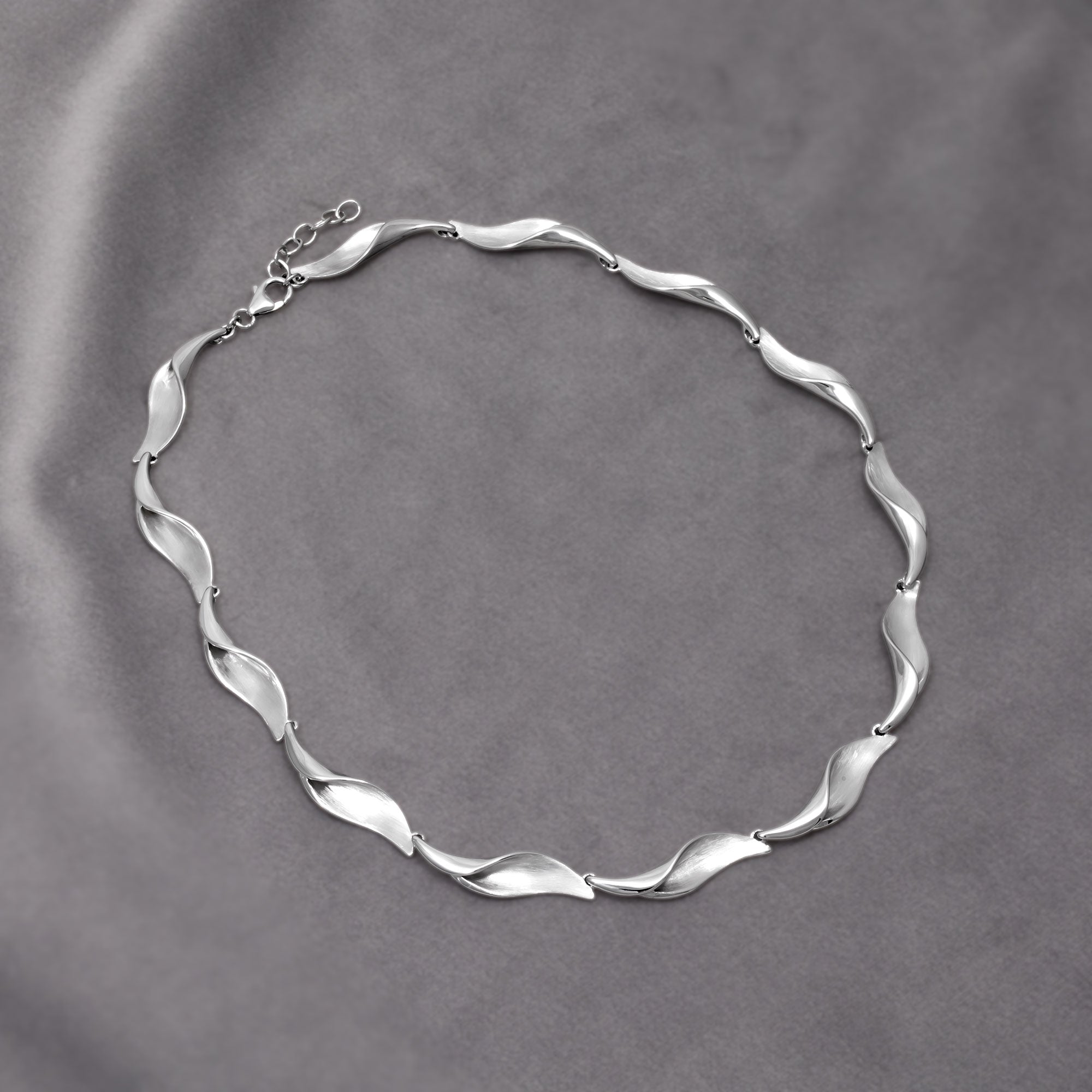 Sterling silver necklace featuring alternating satin and polished links shaped as folded waves, creating a fluid sculptural effect for contemporary silver jewellery. Placed on a grey velvet cloth.