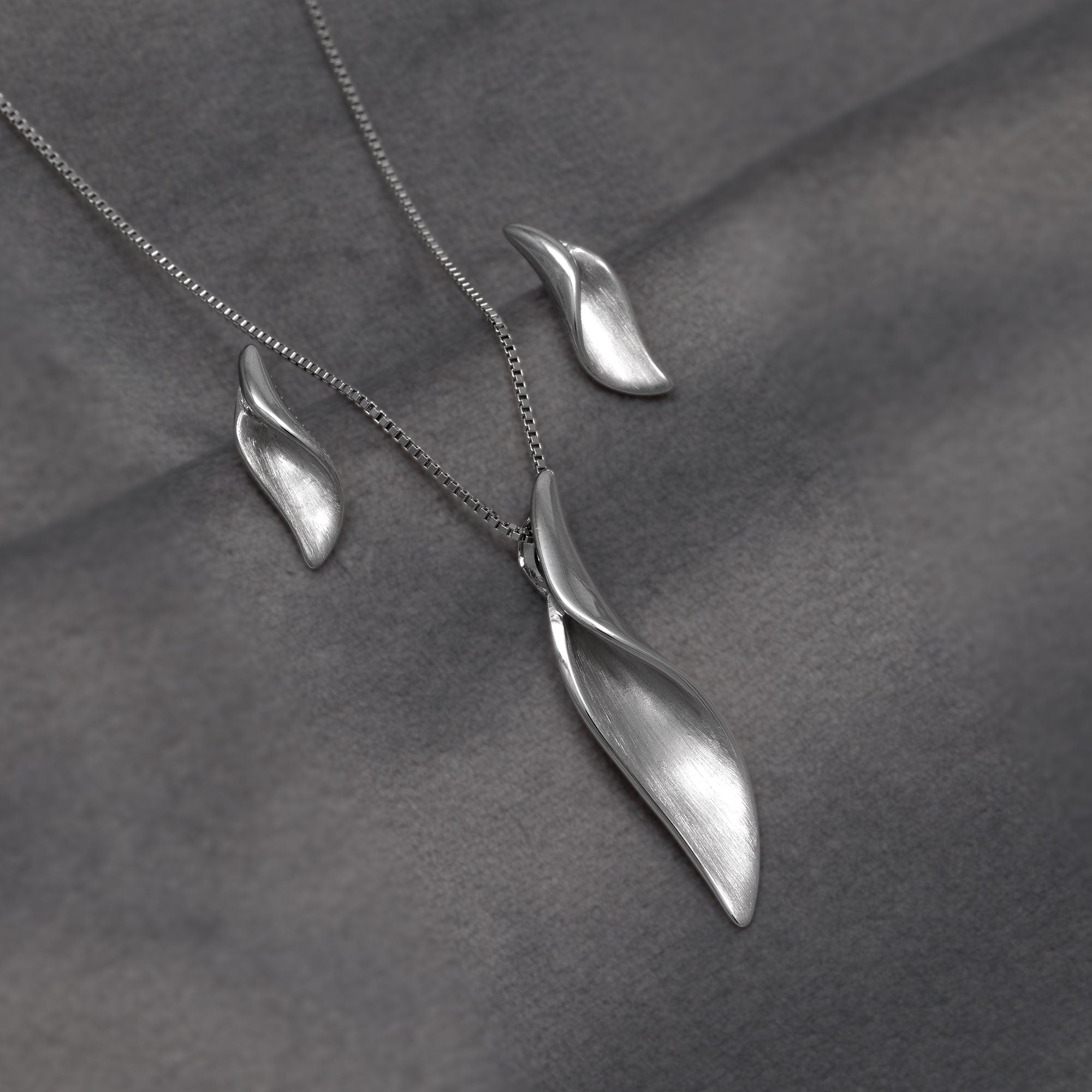 A silver jewellery set featuring a leaf-shaped pendant and a matching leaf stud earring, displayed on a textured dark gray fabric background.