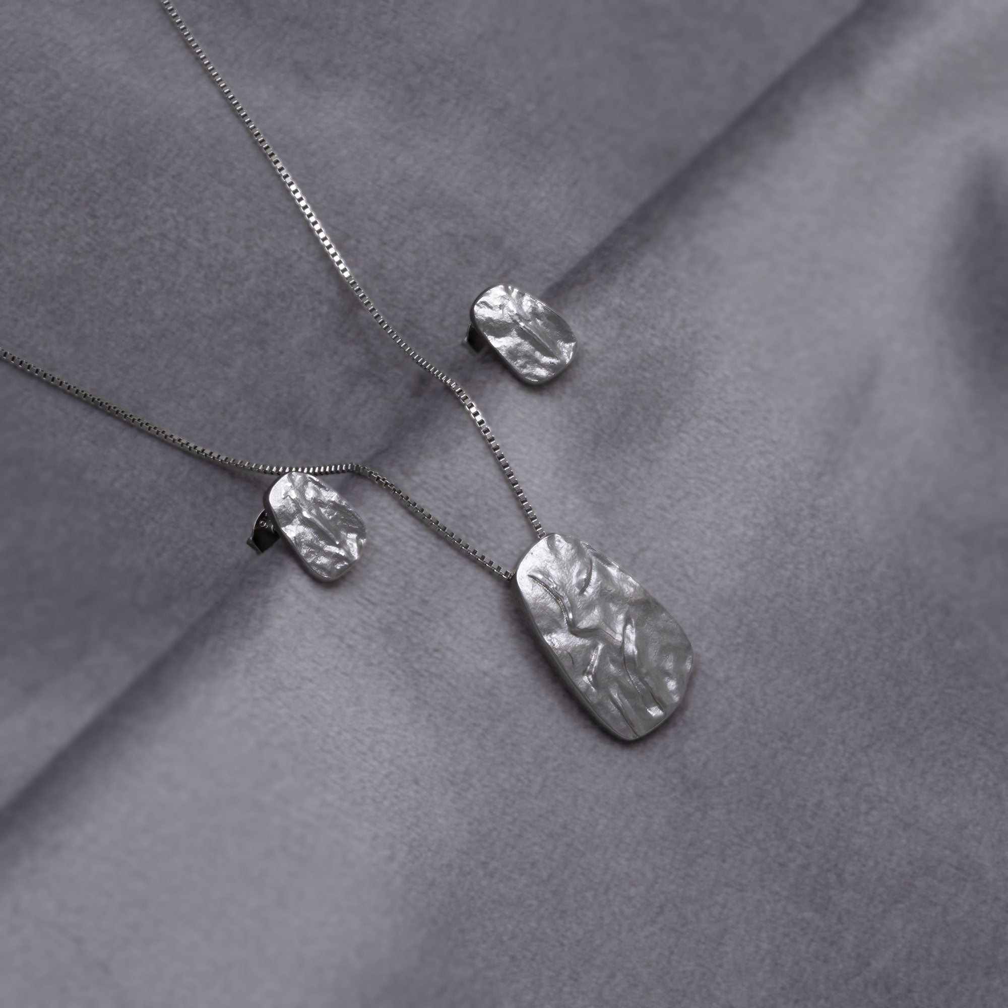 Sterling silver pebble-shaped pendant  and matching stud earrings with a textured, sculptural surface suspended on a delicate chain, reflecting contemporary British silver jewellery design. Placed on a grey velvet cloth.