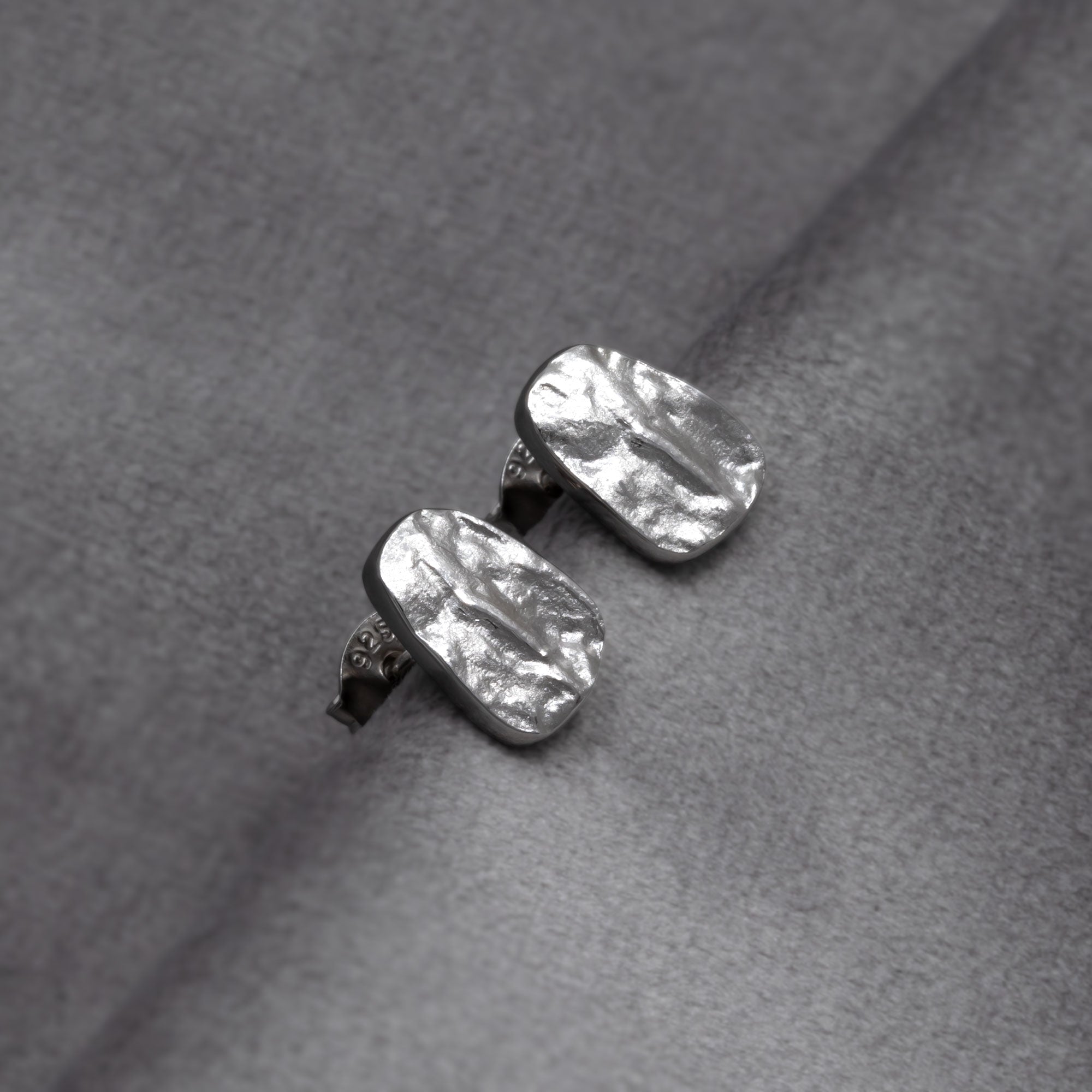 Sterling silver pebble-shaped stud earrings with a textured, sculptural surface suspended on a delicate chain, reflecting contemporary British silver jewellery design. Placed on a grey velvet cloth.
