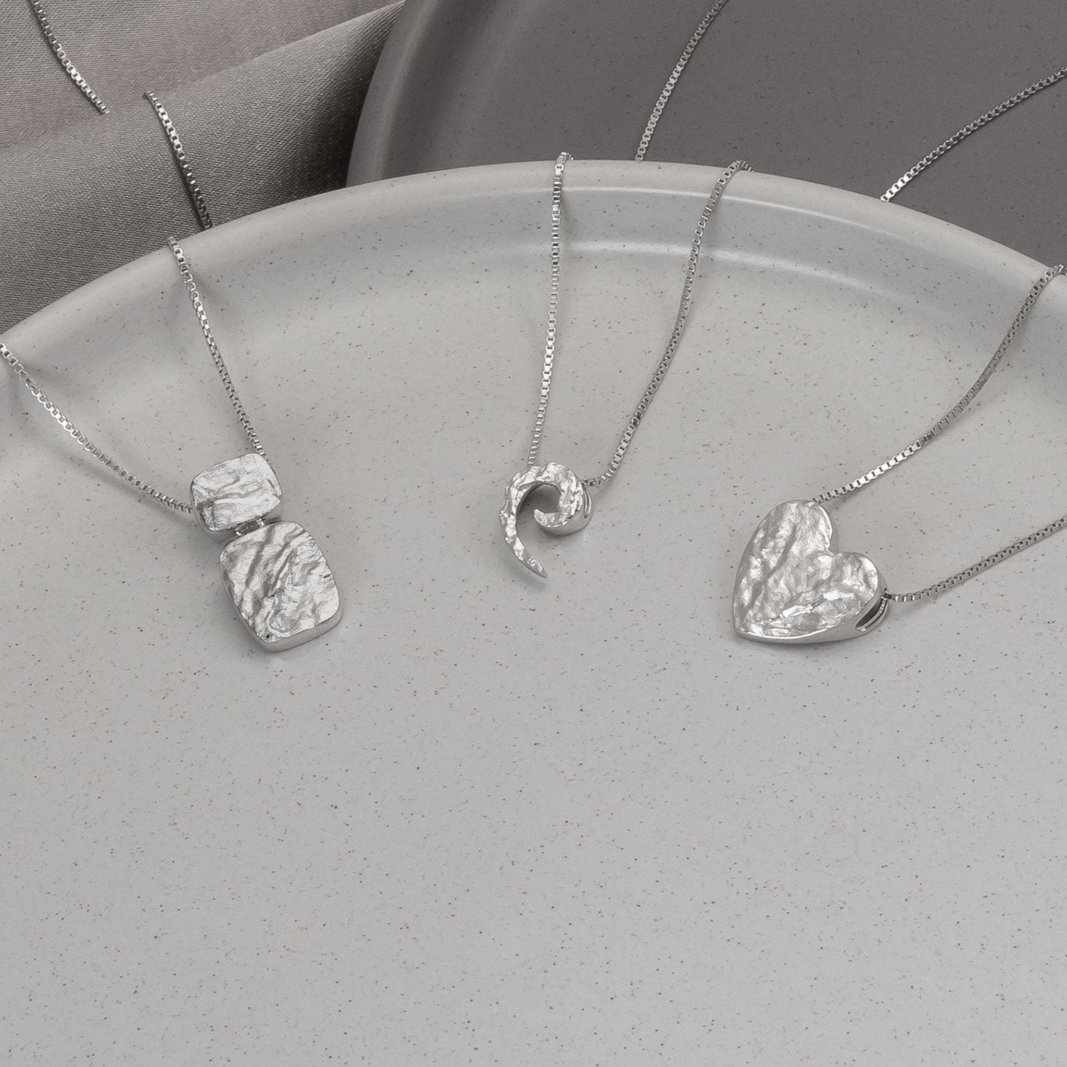 Collection of sterling silver textured pendants including a sculptural heart, organic swirl, and geometric shapes, each suspended on delicate fine silver chains.