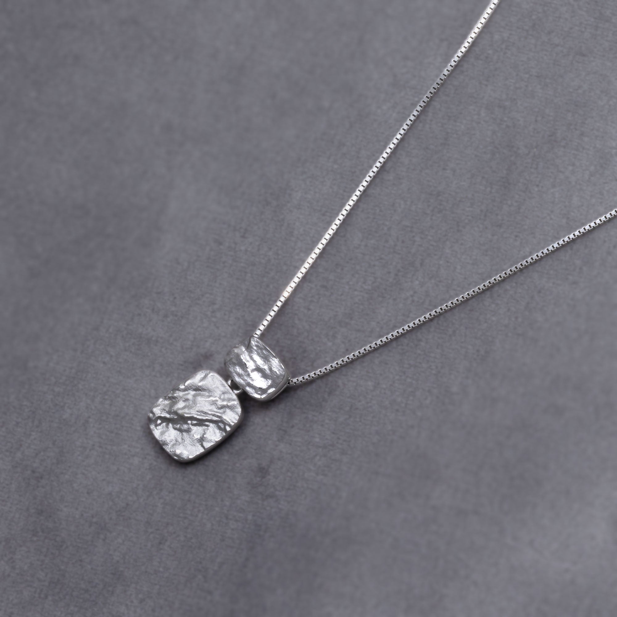 Sterling silver pendant featuring two textured rectangular elements with an organic, sculptural finish on a fine chain, embodying refined contemporary silver jewellery. Placed on a grey velvet cloth.