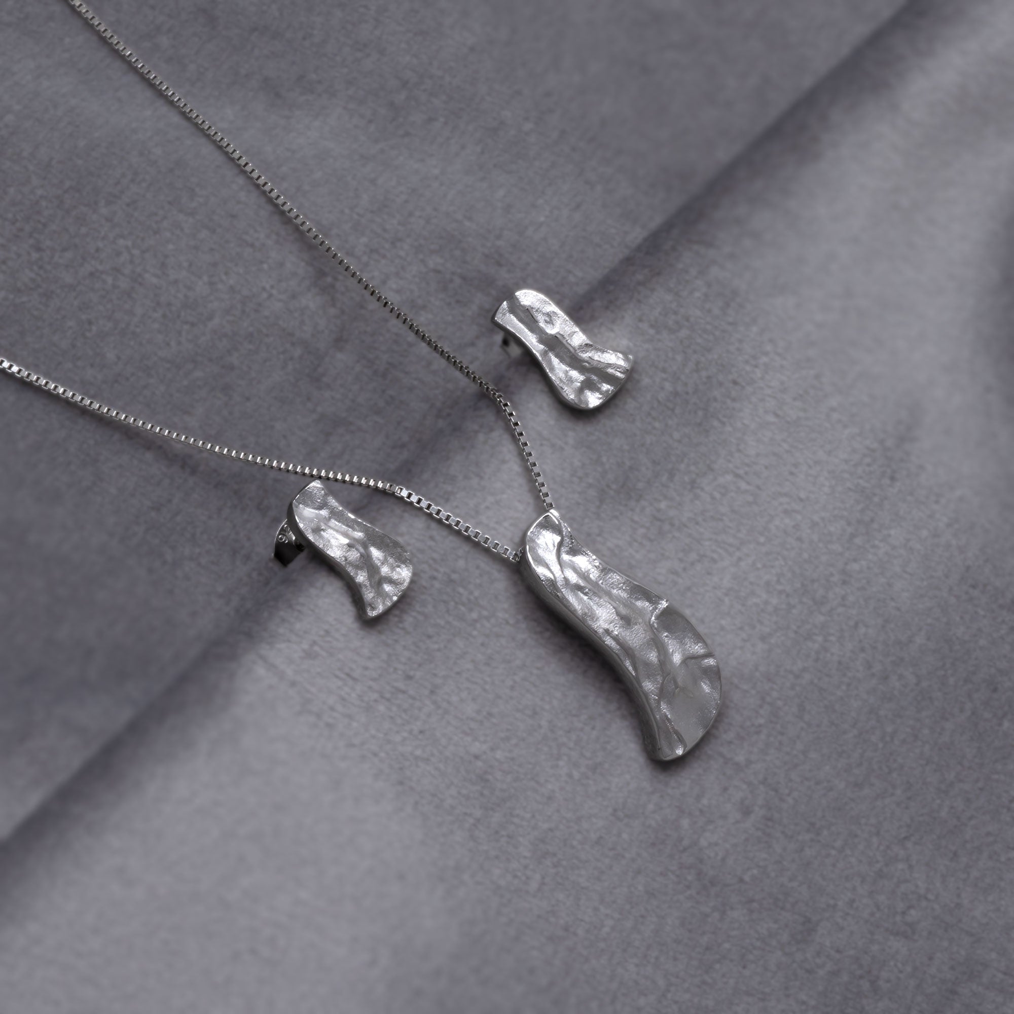 Contemporary sterling silver pendant and matching stud earrings with an organic, asymmetrical shape and hammered texture, suspended on a delicate box chain for a modern, sculptural look. Placed on a grey velvet cloth. 