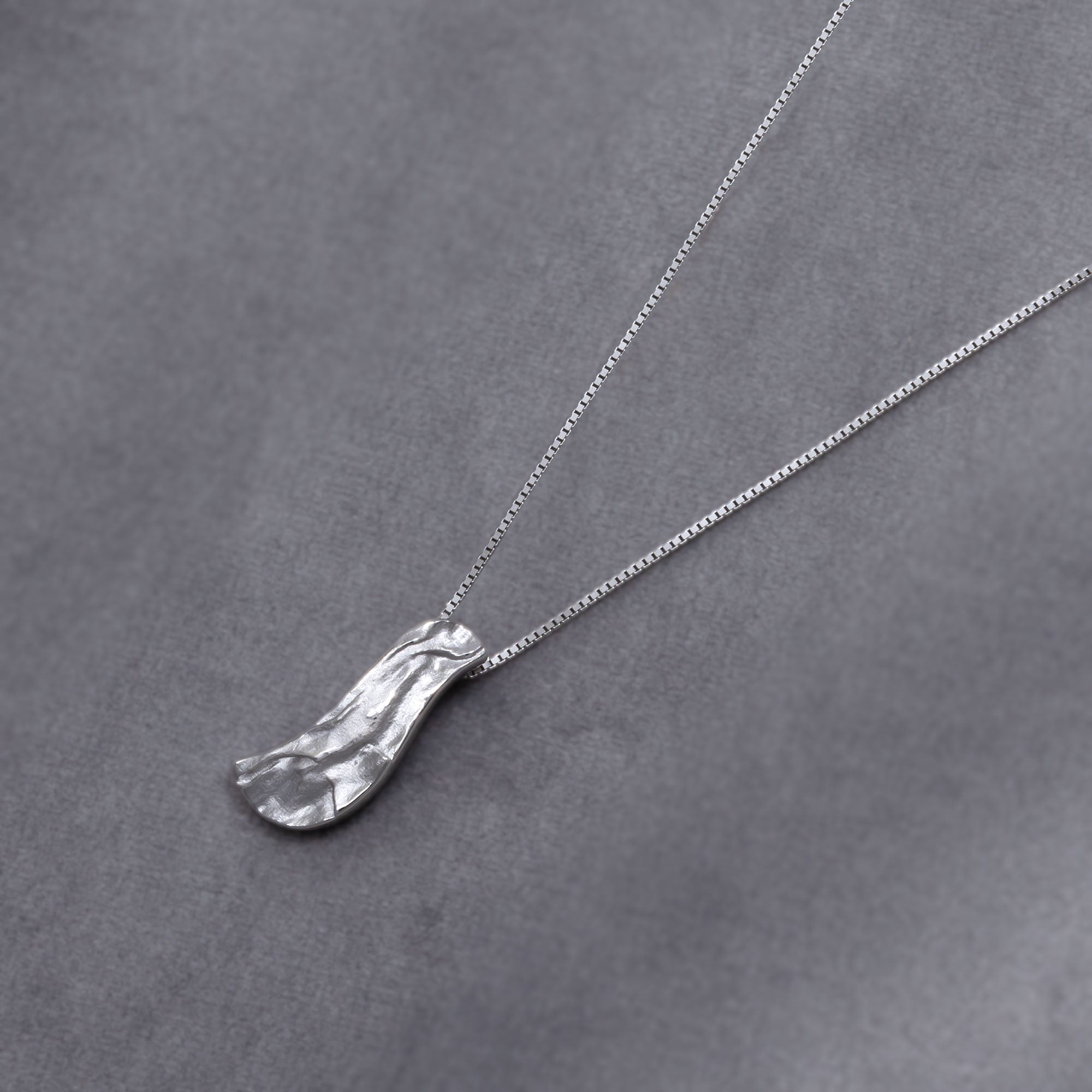 Contemporary sterling silver pendant with an organic, asymmetrical shape and hammered texture, suspended on a delicate box chain for a modern, sculptural look. Placed on a grey velvet cloth. 