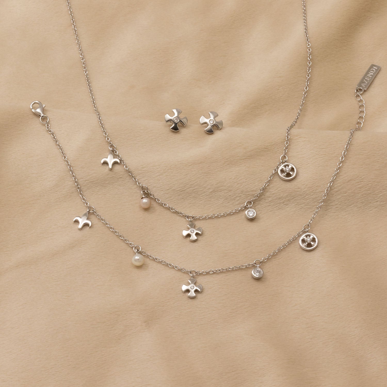 Two sterling silver charm bracelets featuring fleur de lis, cross, and circular motifs, accented with delicate pearls and small stones, displayed on a soft beige background.