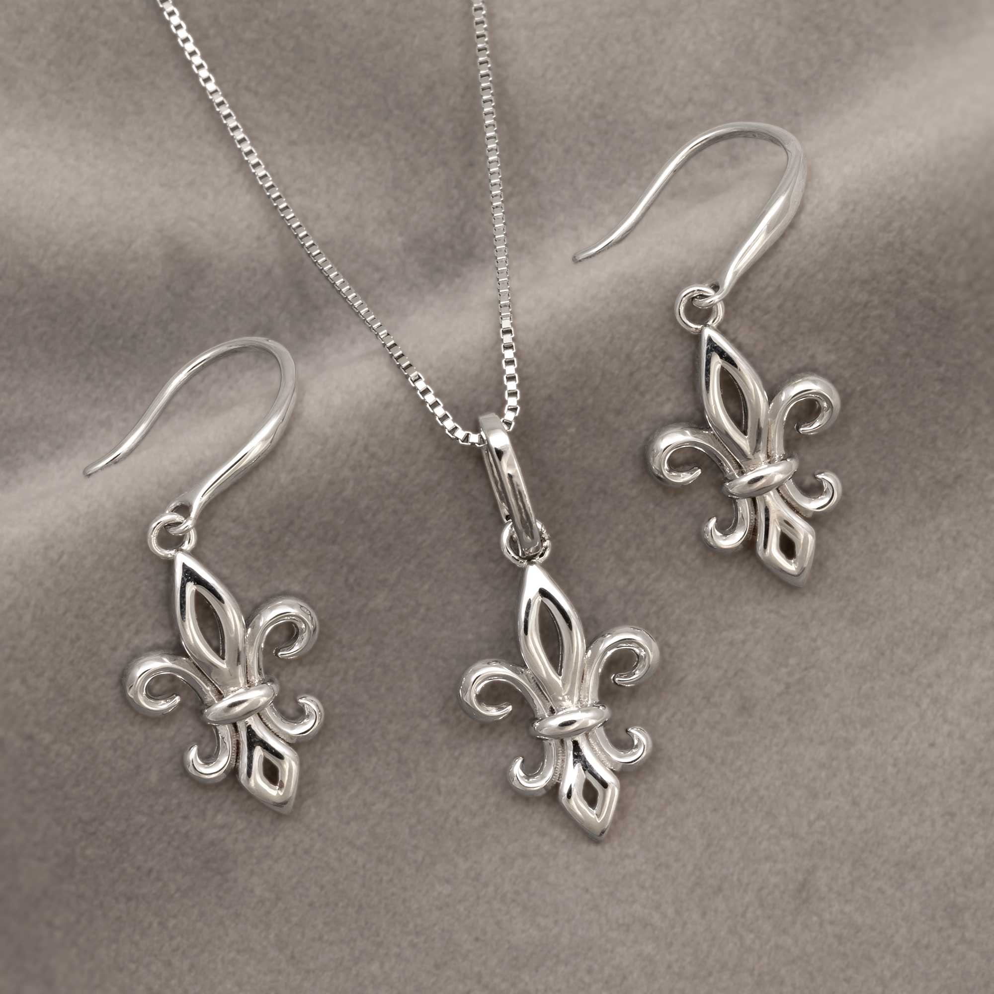Sterling silver pendant and drop earrings featuring a sculptural fleur de lis design suspended from a delicate silver chain, embodying timeless elegance and expert craftsmanship. Placed on grey velvet cloth.