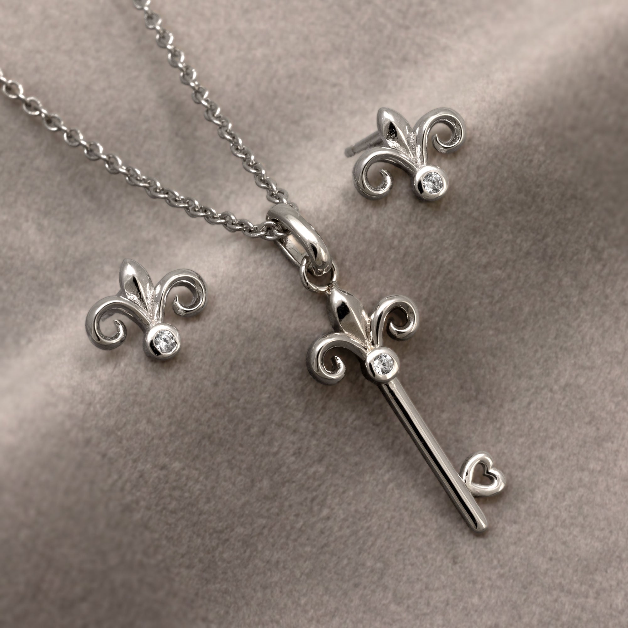 Delicate sterling silver necklace and stud earrings featuring a sculptural fleur de lis key pendant suspended from a fine chain, showcasing refined British design by Azendi. Placed on a grey velvet cloth.