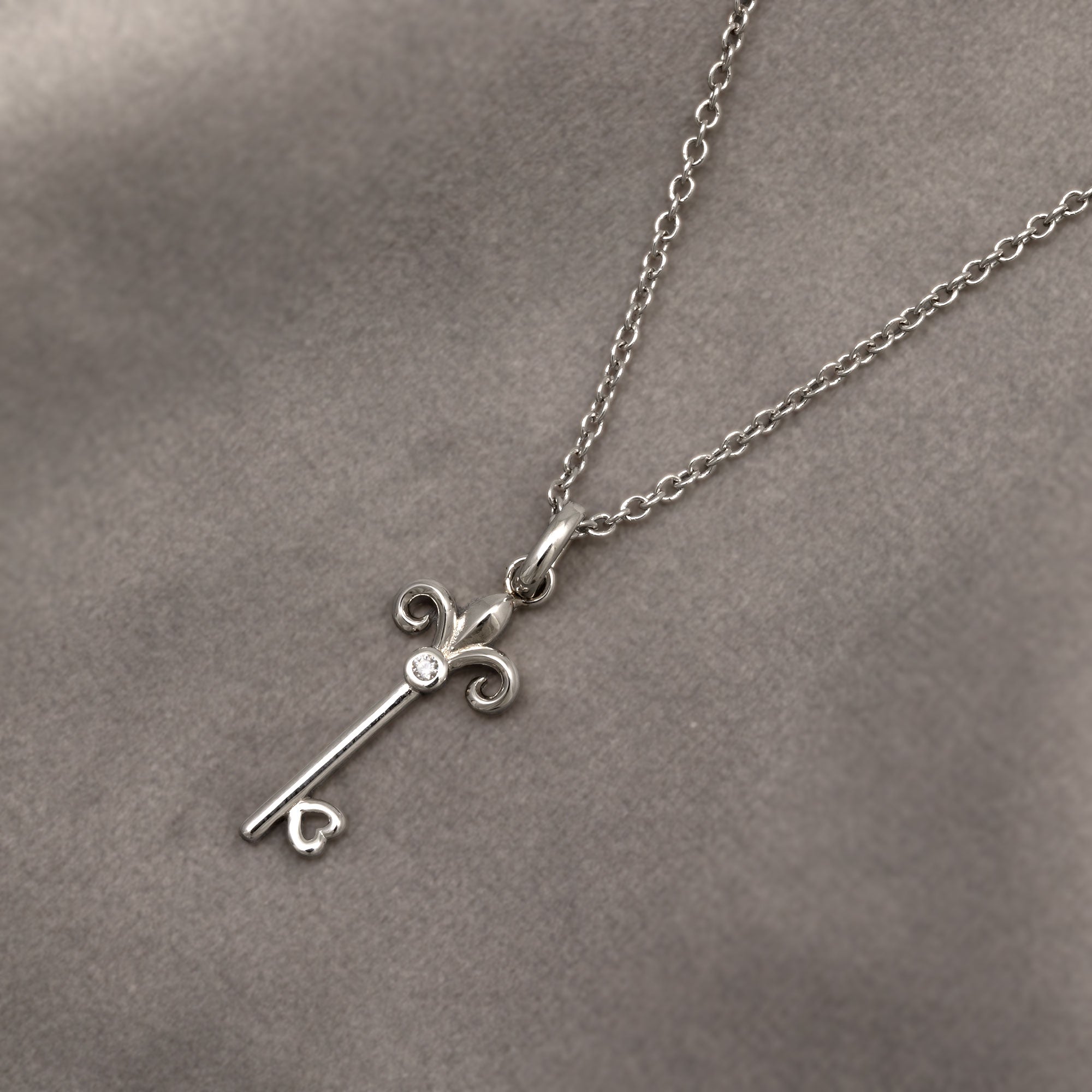 Delicate sterling silver necklace featuring a sculptural fleur de lis key pendant suspended from a fine chain, showcasing refined British design by Azendi. Placed on a grey velvet cloth.
