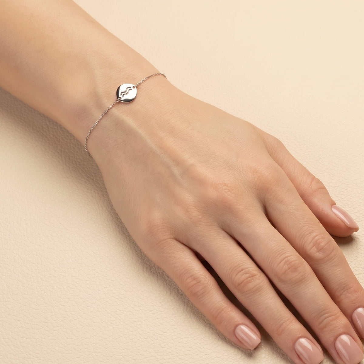 Delicate sterling silver bracelet featuring a polished Aquarius symbol charm on a fine adjustable chain, styled on the wrist against a cream background.
