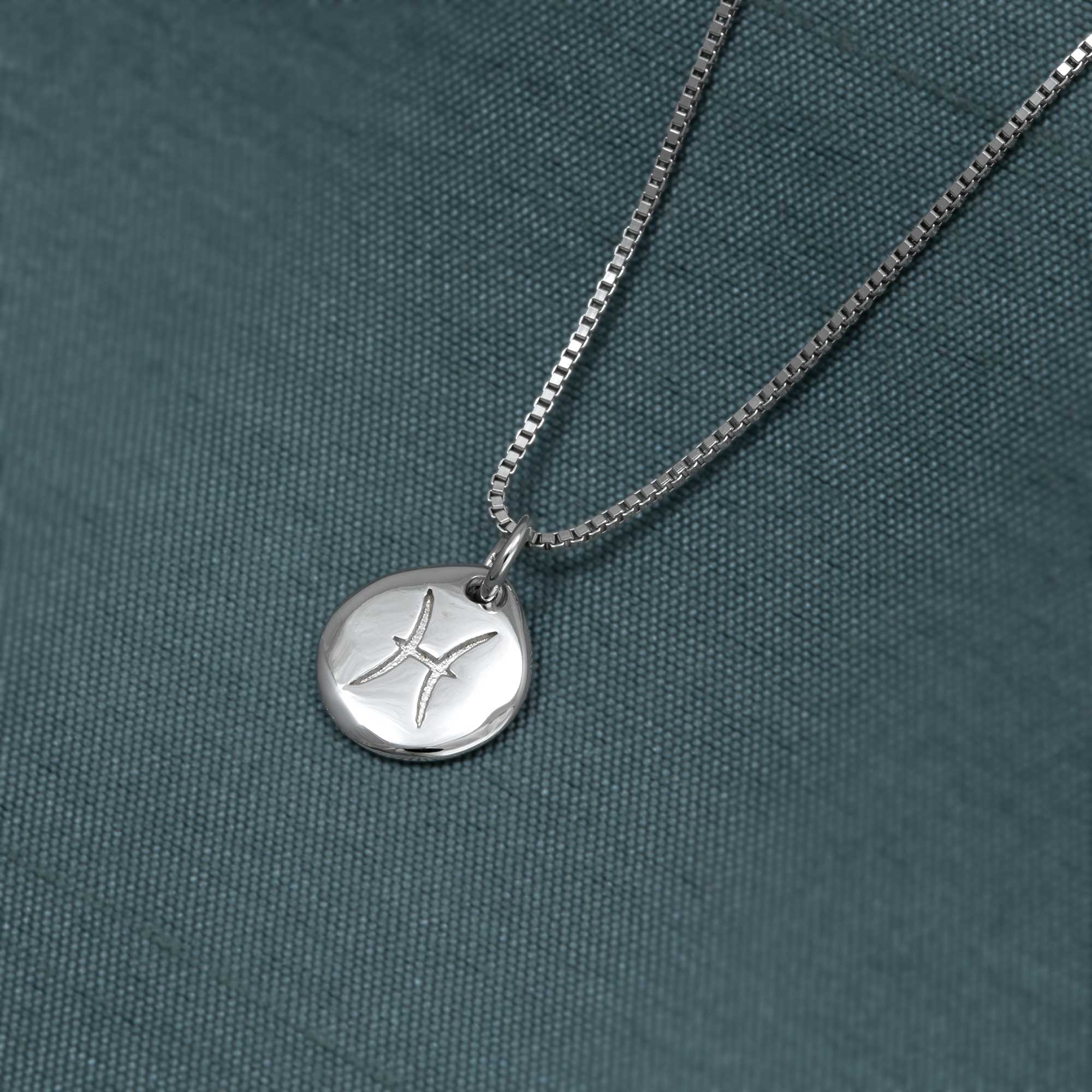 Sterling silver pendant featuring a sculptural Pisces zodiac symbol, suspended from a delicate box chain and displayed against a textured dark teal fabric background.