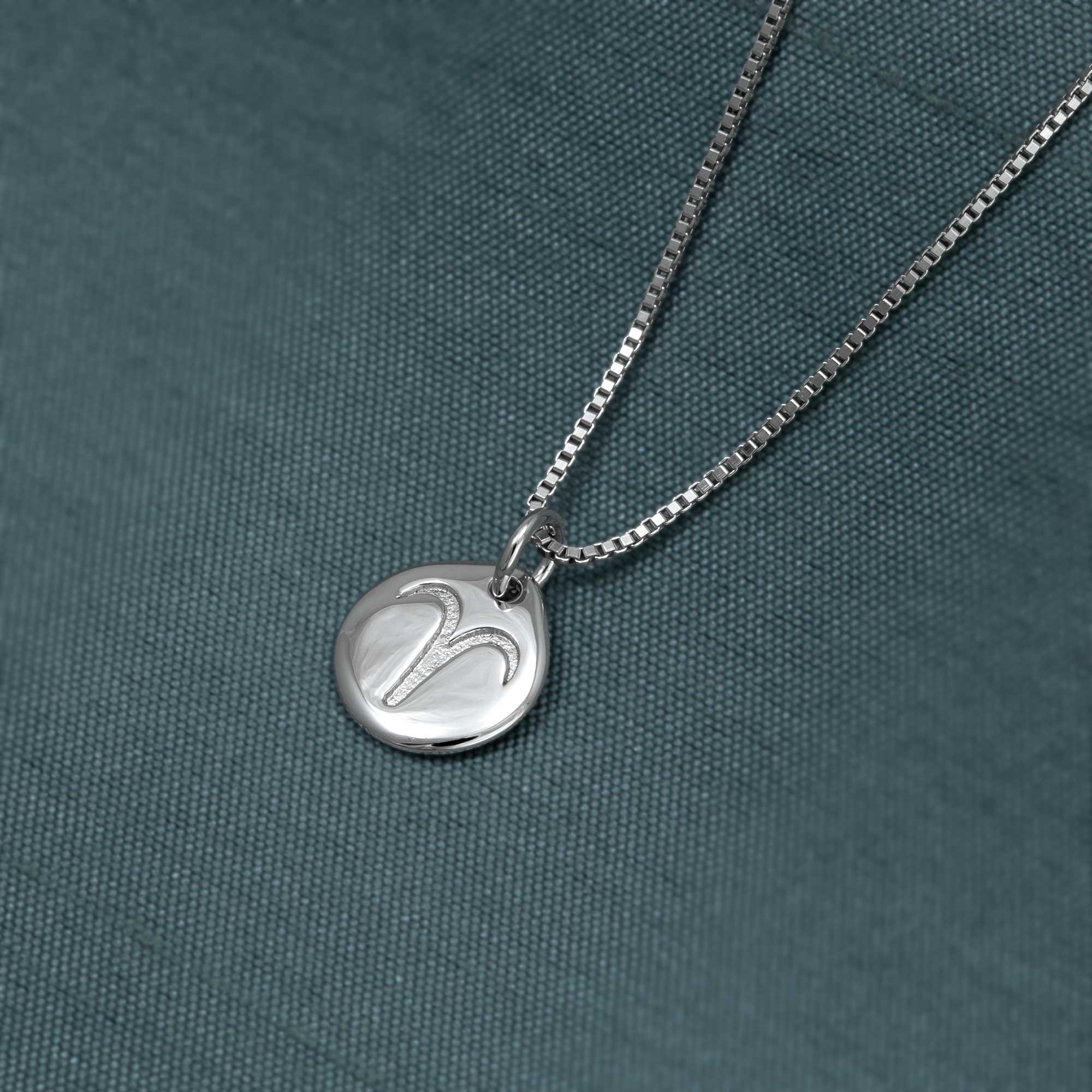 Sterling silver pendant necklace featuring a polished circular charm engraved with the Aries zodiac symbol, displayed against a textured dark green fabric background.