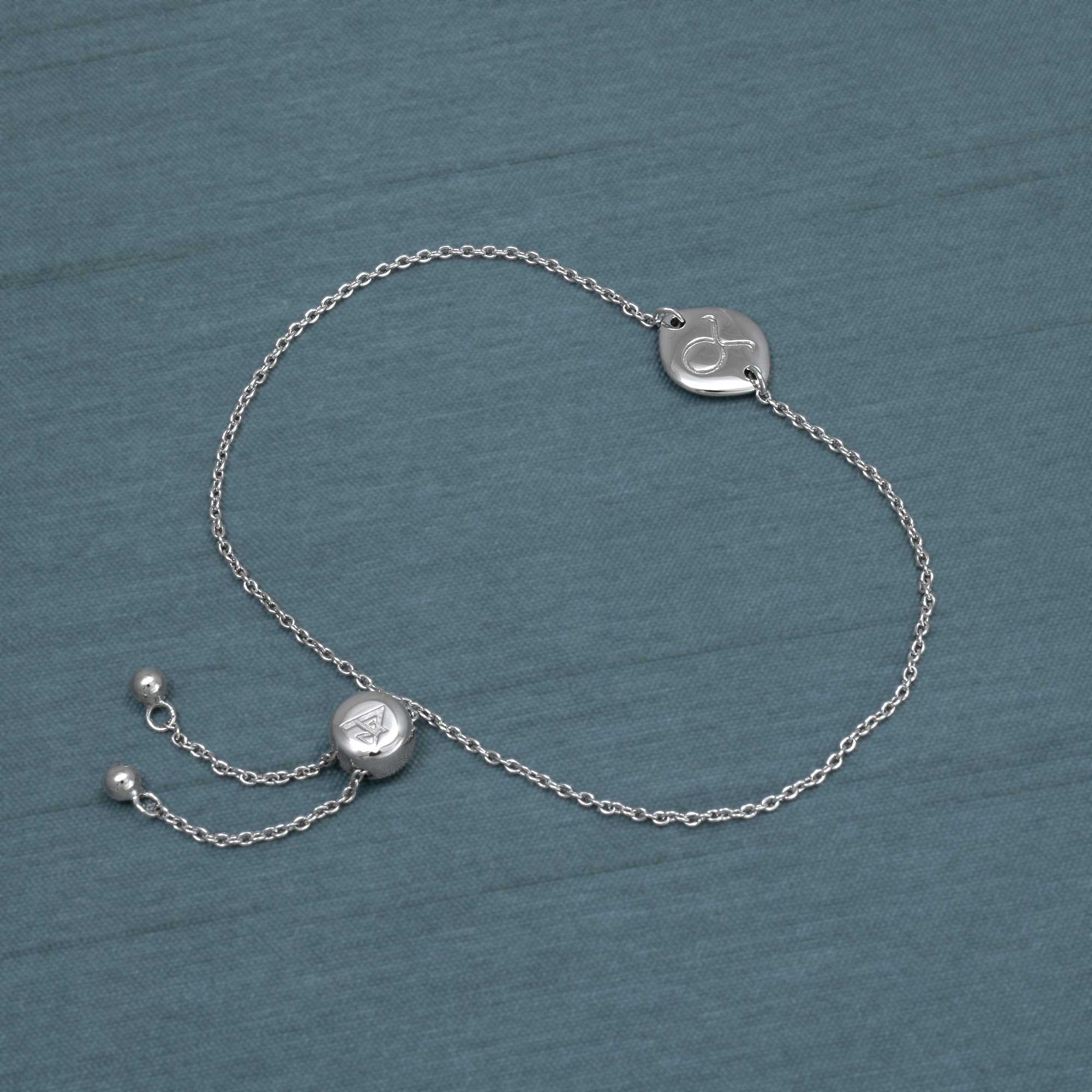 Delicate sterling silver adjustable bracelet featuring a Taurus zodiac charm with a sliding bead closure, crafted with fine chain links on a muted blue background.