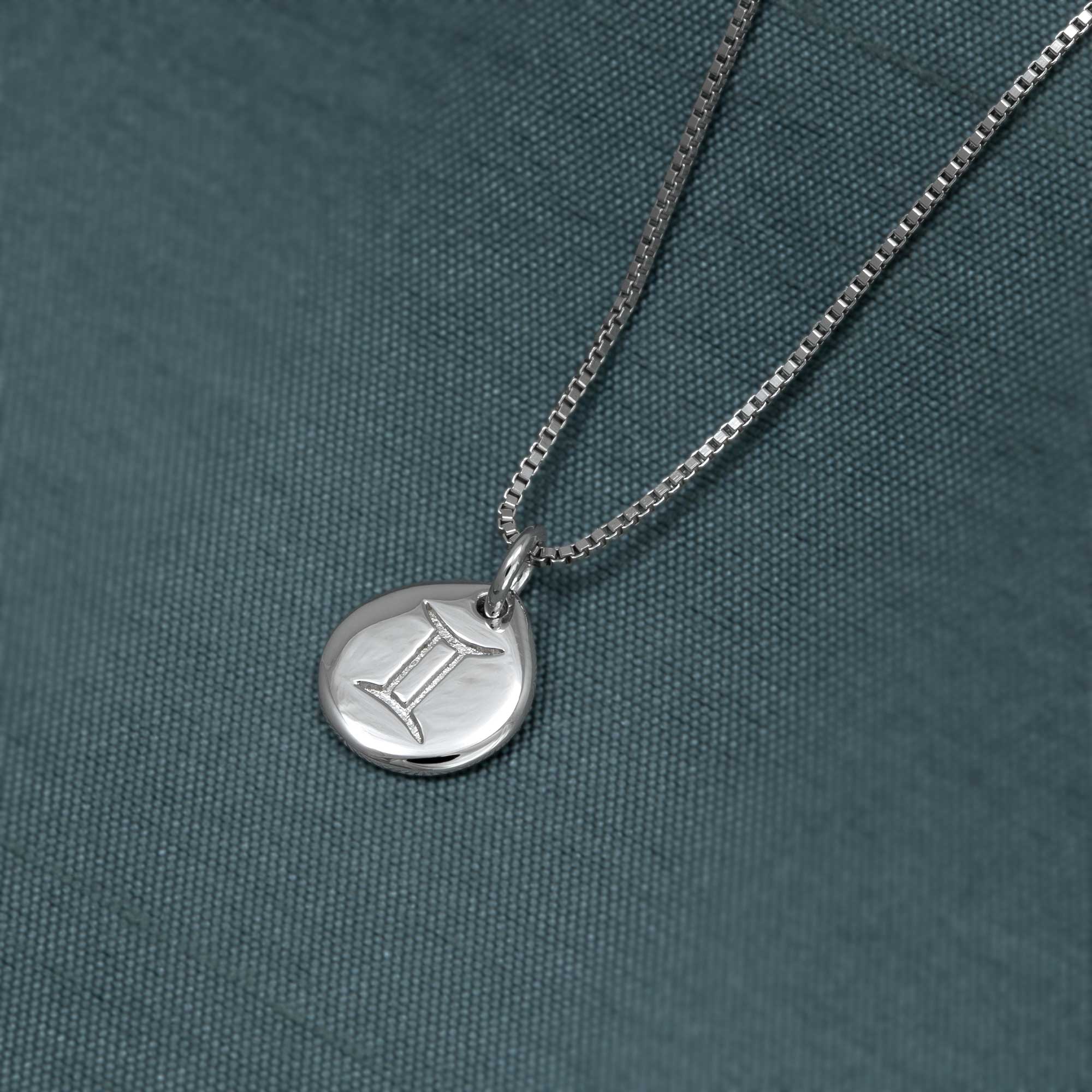Sterling silver pendant featuring an engraved Gemini zodiac symbol, suspended from a delicate box chain, displayed on a textured blue fabric background.