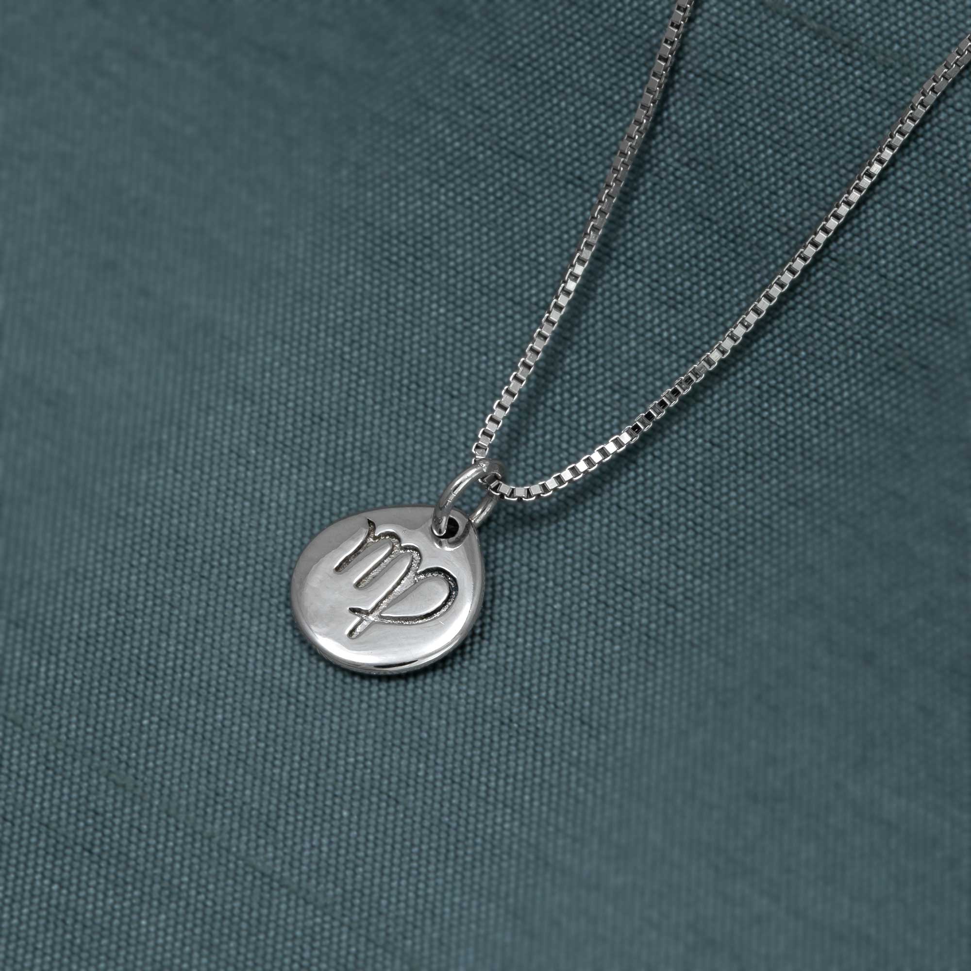 Sterling silver pendant featuring a sculptural Virgo zodiac symbol engraved on a polished circular disc, suspended from a sleek, modern box chain against a textured fabric background.