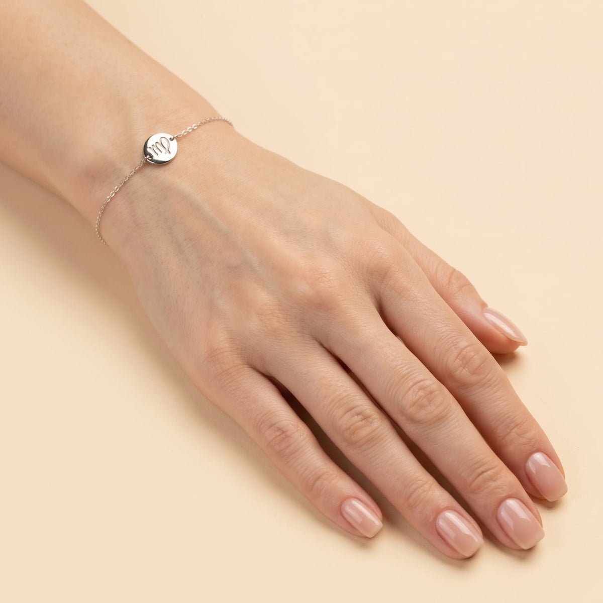 Delicate sterling silver adjustable bracelet featuring a polished round charm engraved with the Virgo zodiac symbol, showcased on a wrist against a cream background.