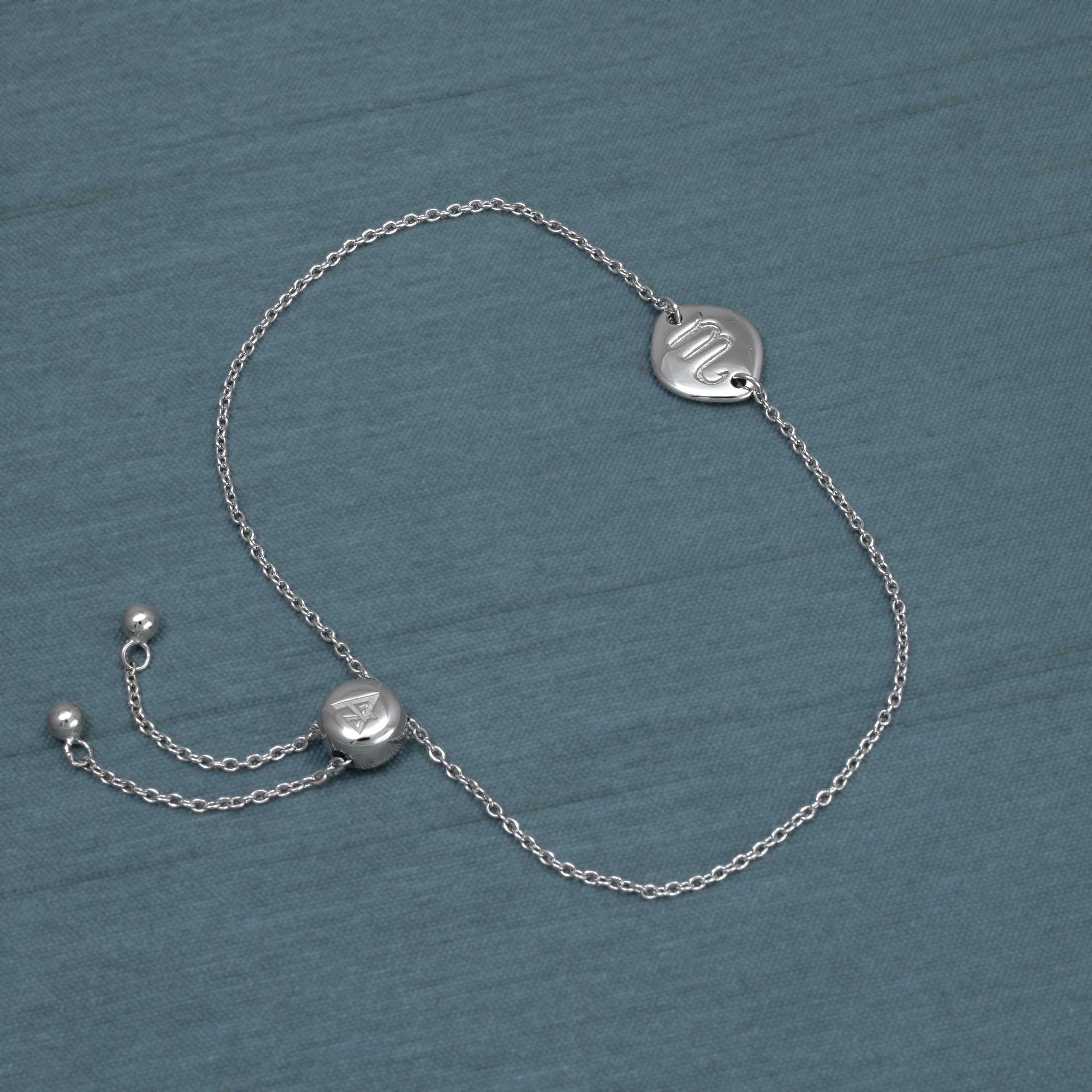 Delicate sterling silver adjustable bracelet featuring a Scorpio zodiac disc charm and a sliding bead closure, styled on a soft blue fabric background.