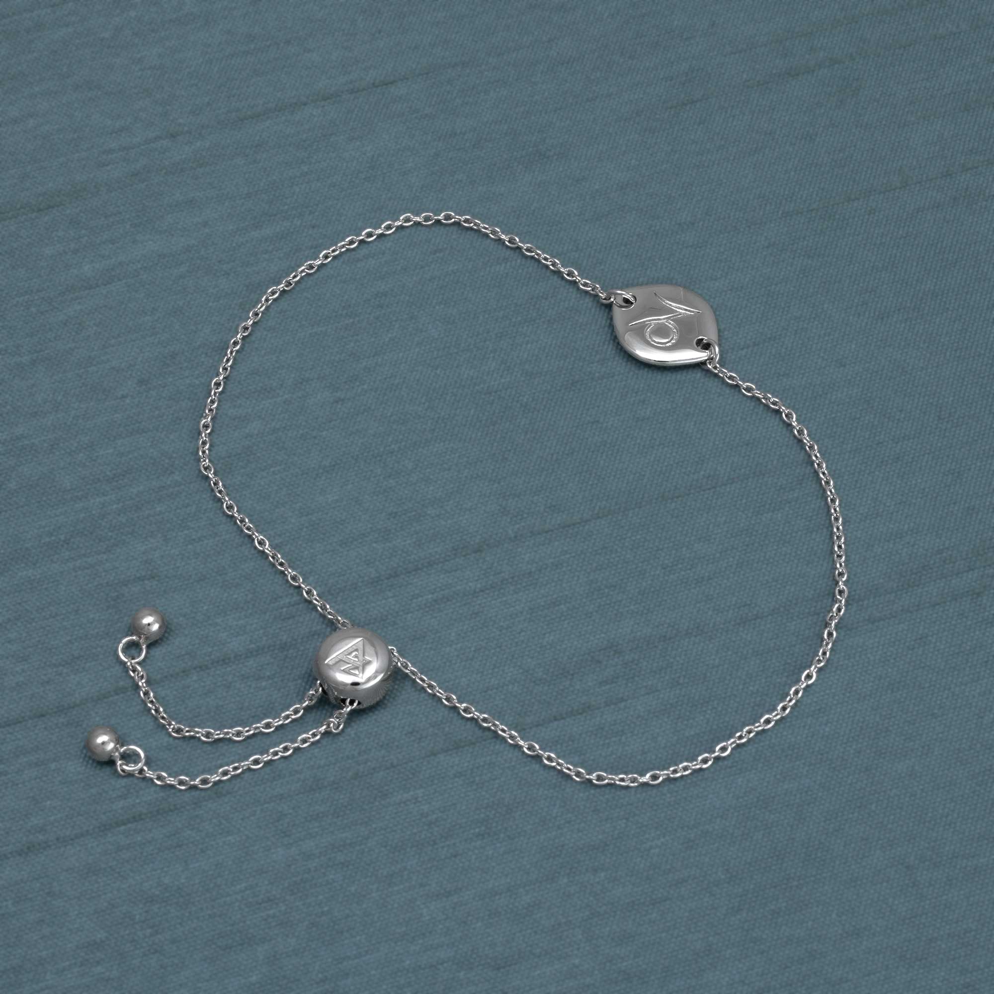 Delicate sterling silver adjustable bracelet featuring a Capricorn zodiac charm with a smooth polished finish, styled on a muted blue fabric background.