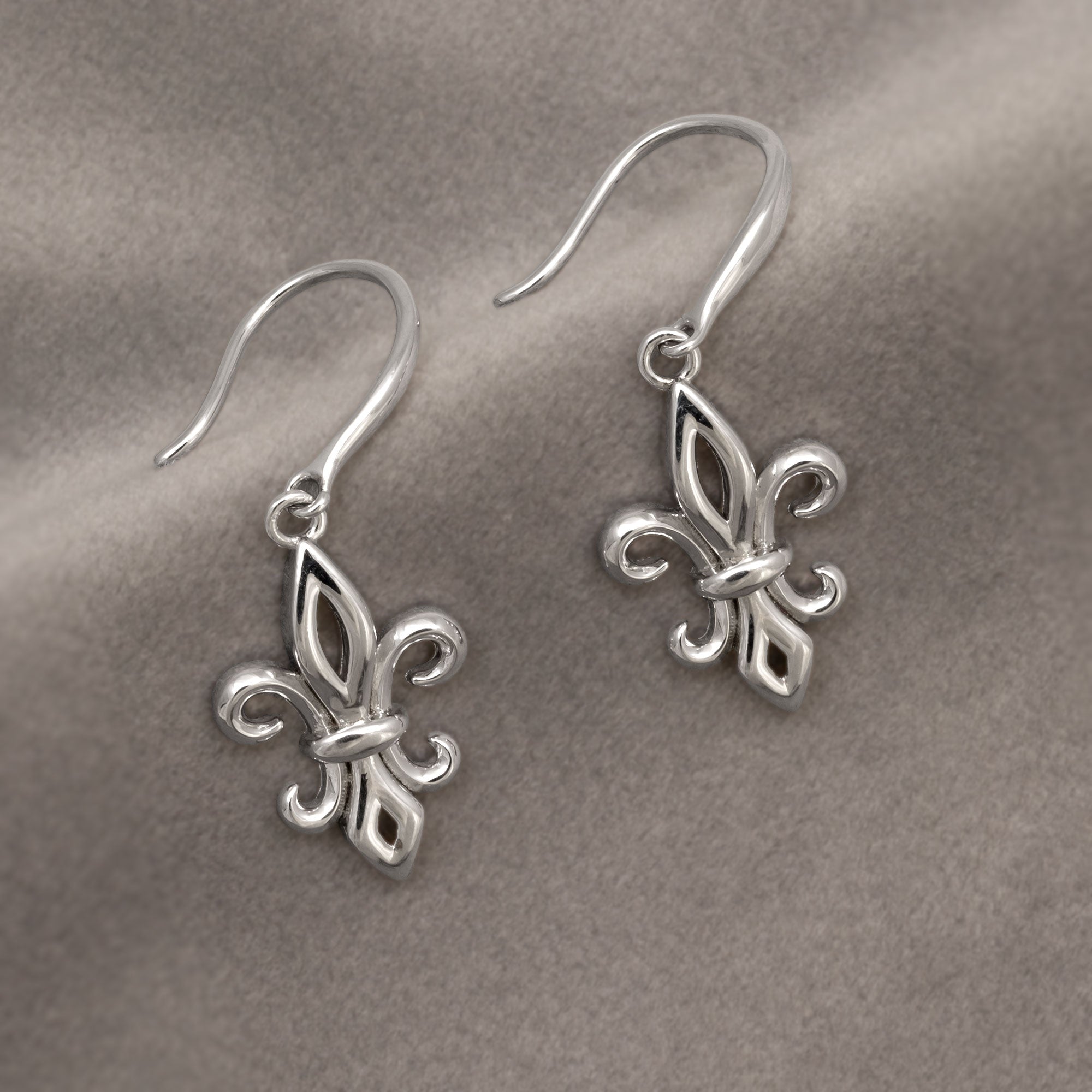 Pair of sterling silver drop earrings featuring elegant fleur de lis charms suspended from curved hooks, showcasing a polished and sculptural design. Placed on grey velvet cloth.