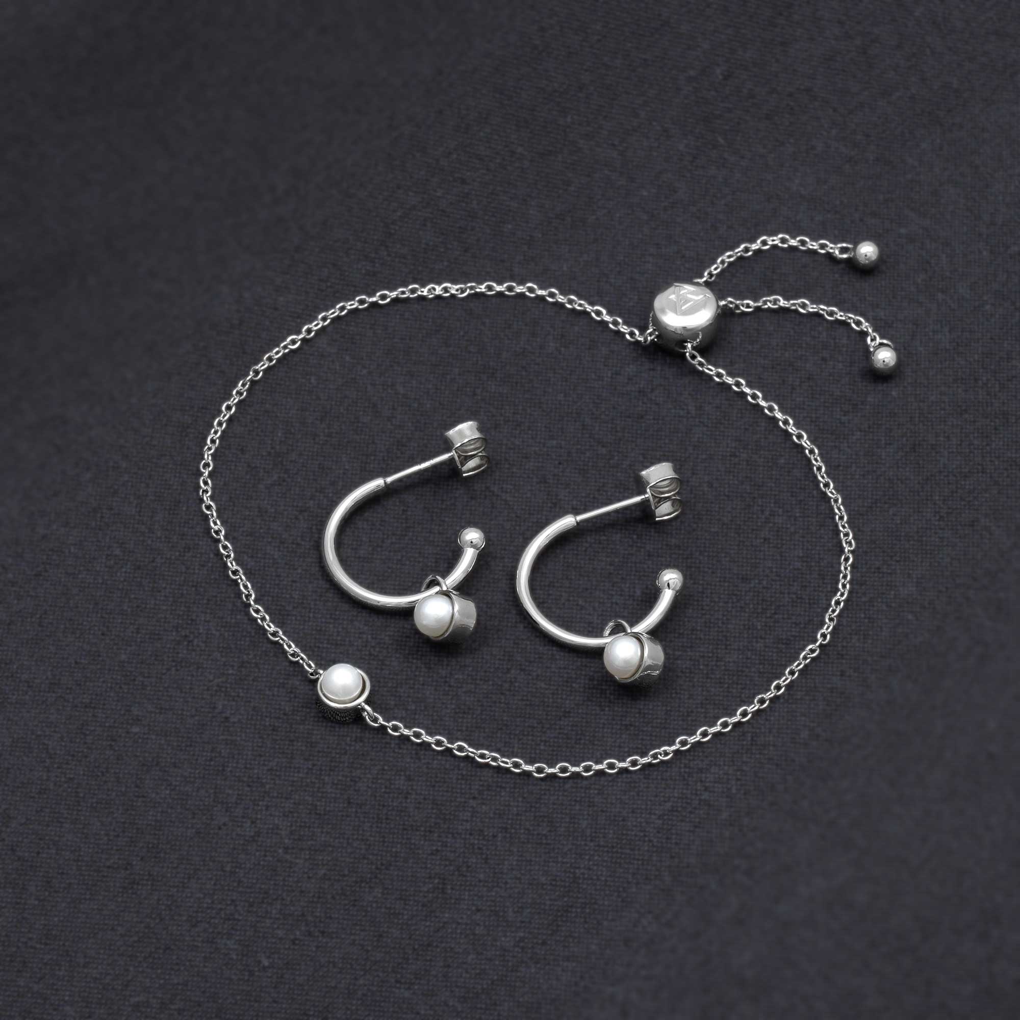 Sterling silver hoop earrings featuring freshwater pearls paired with a delicate chain bracelet, all displayed on a smooth dark fabric backdrop.