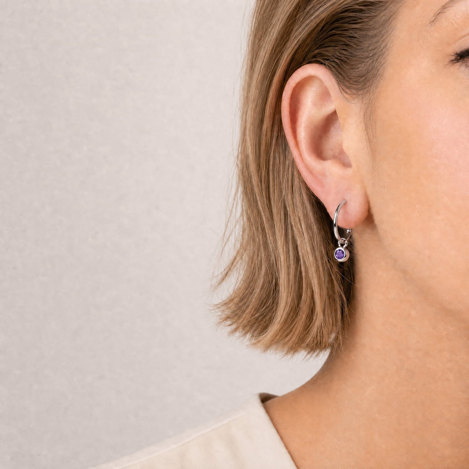 Model wears sterling silver hoop earrings featuring a faceted amethyst birthstone charm, combining minimalist design with expert British craftsmanship for a subtle, elegant accessory.