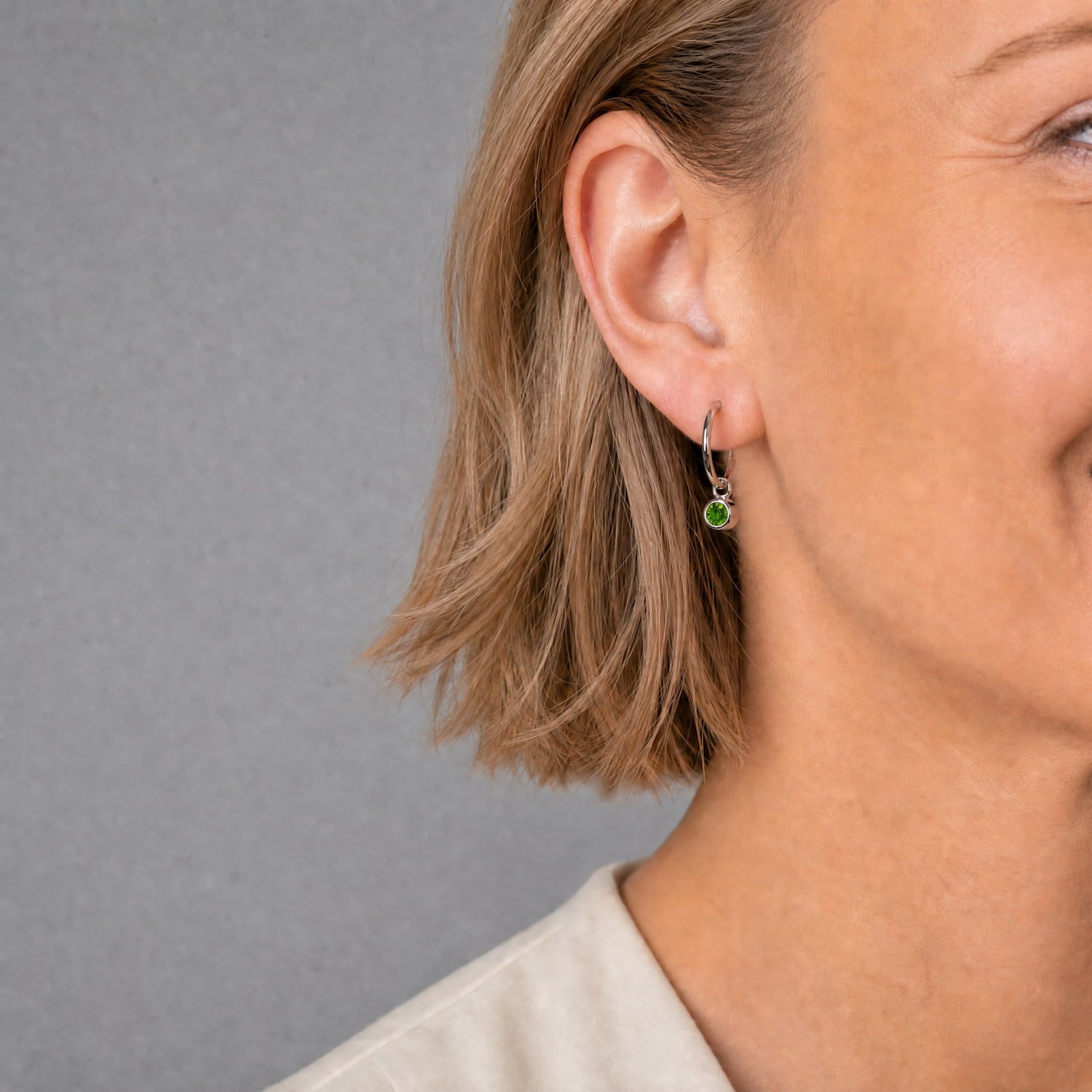 Delicate sterling silver hoop earrings featuring a vibrant Siberian emerald birthstone, highlighted by a sleek, contemporary design worn by a smiling woman.