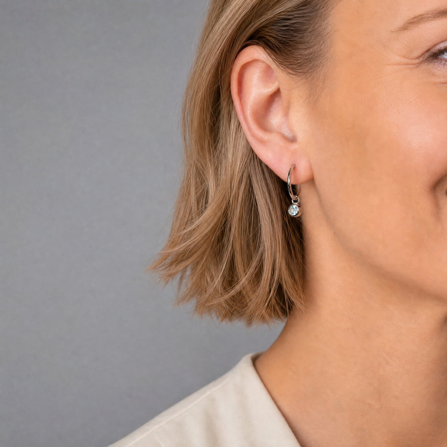Model wears delicate sterling silver hoop earrings featuring round blue topaz birthstone charms, showcasing a refined design with expert British craftsmanship from Azendi.