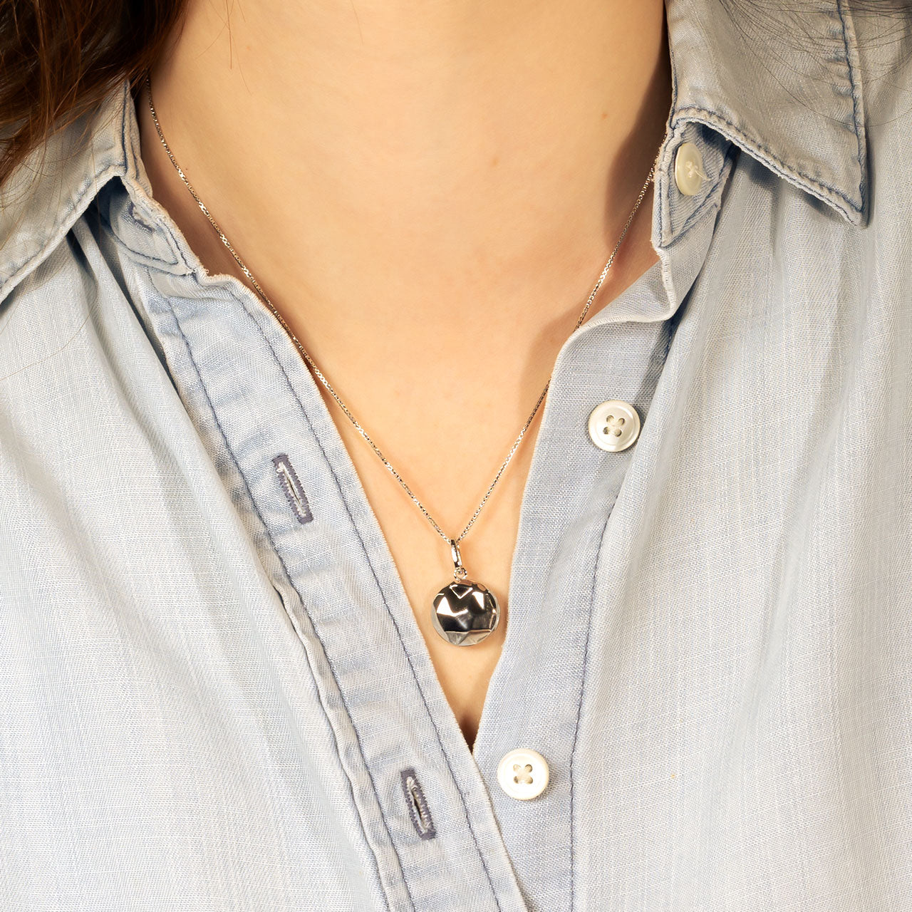 Close-up of a faceted sterling silver locket pendant hanging from a delicate adjustable chain, styled with a light blue button-down shirt for a refined look.
