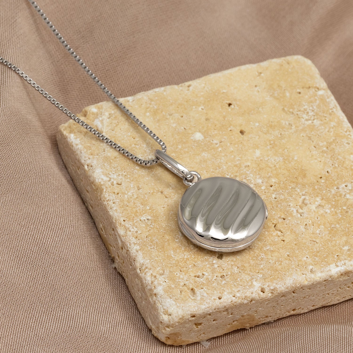 Polished sterling silver locket pendant with subtle twisted detailing, suspended from an adjustable fine chain, displayed on a textured natural stone surface.