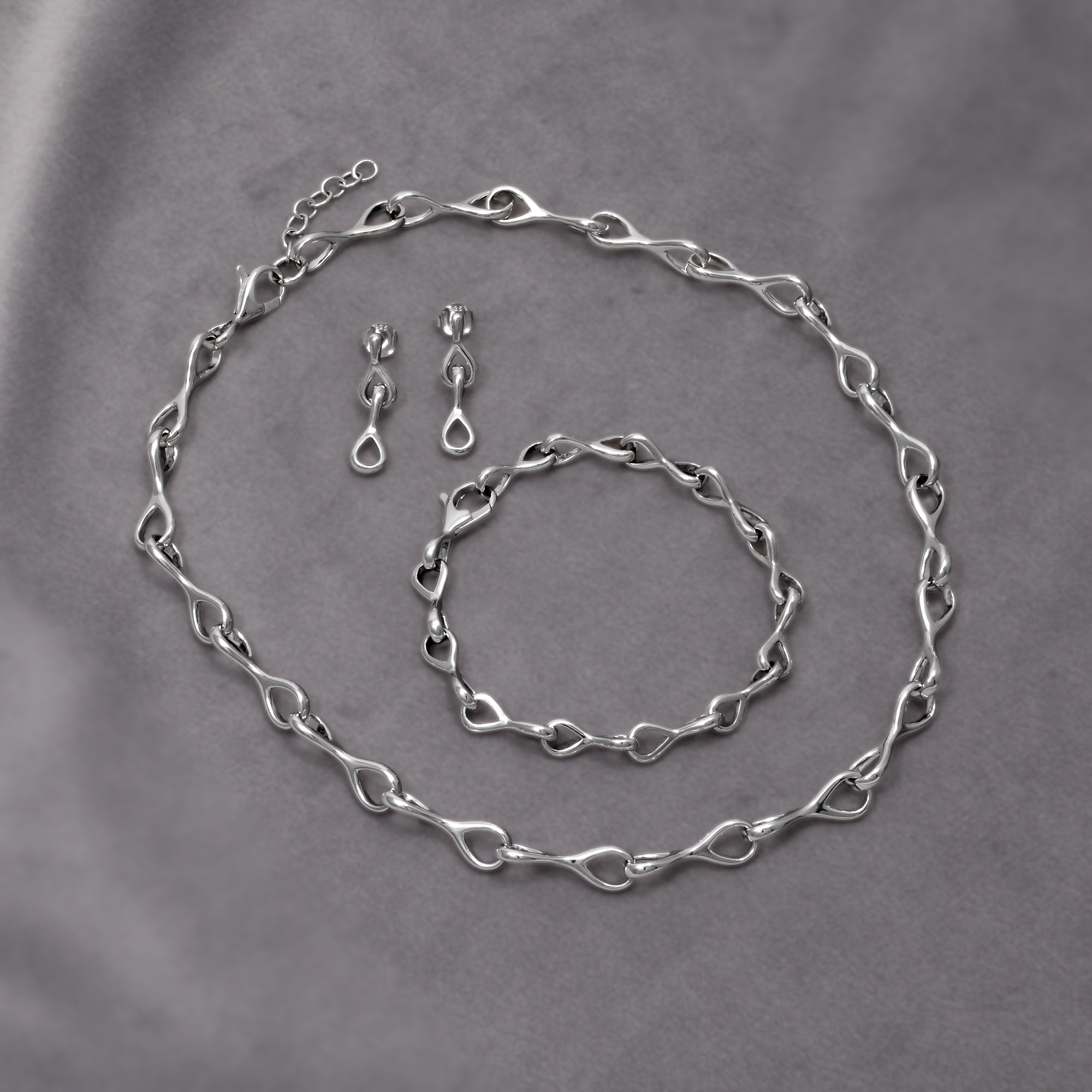 Set of polished sterling silver necklace, bracelet and drop earrings featuring interlocking teardrop links with smooth, flowing contours for a modern, sculptural expression in silver jewellery. Placed on a grey velvet cloth.
